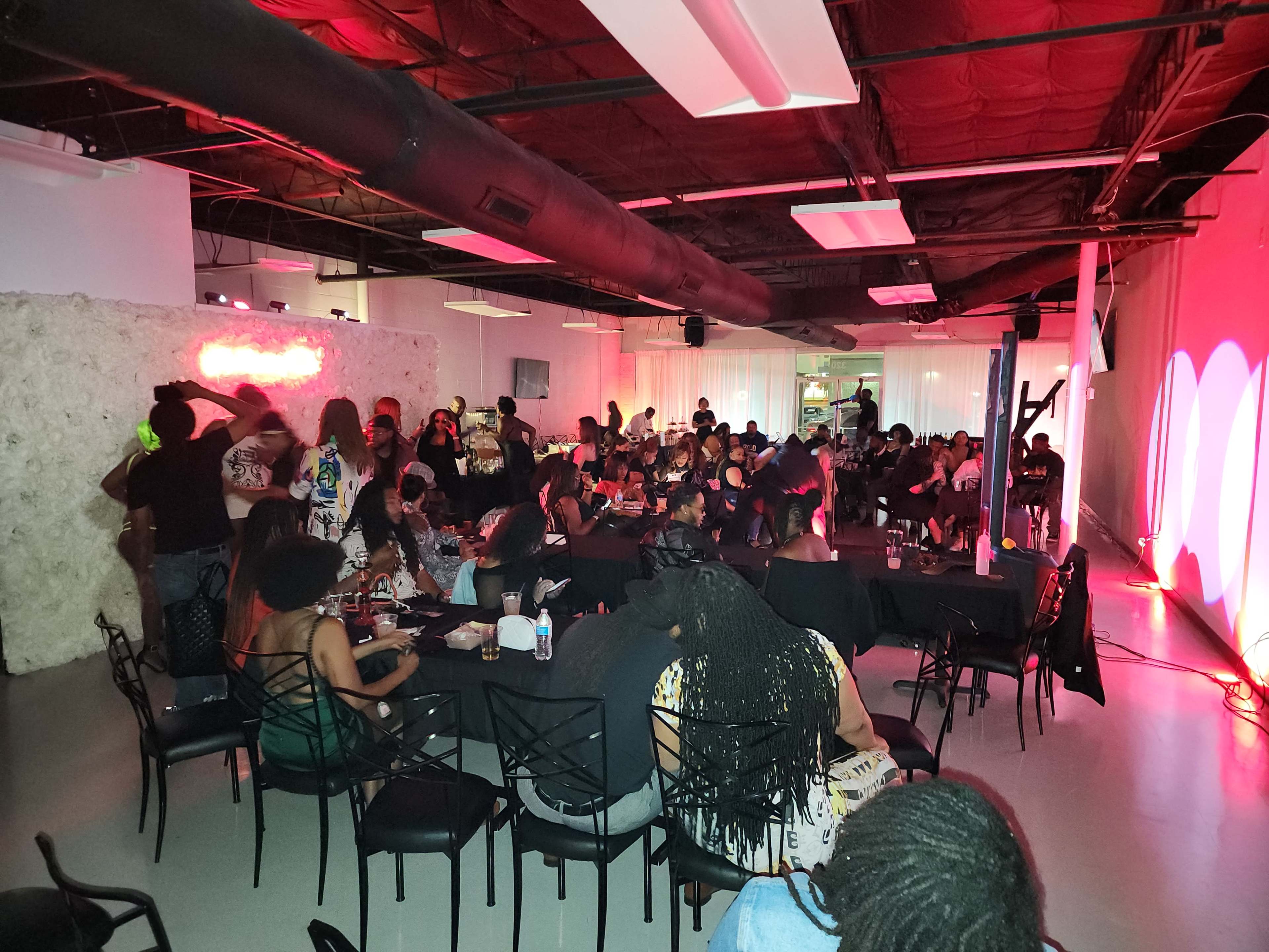 A crowded venue filled with people seated at black tables, some socializing, and others enjoying a performance under colorful lighting.
