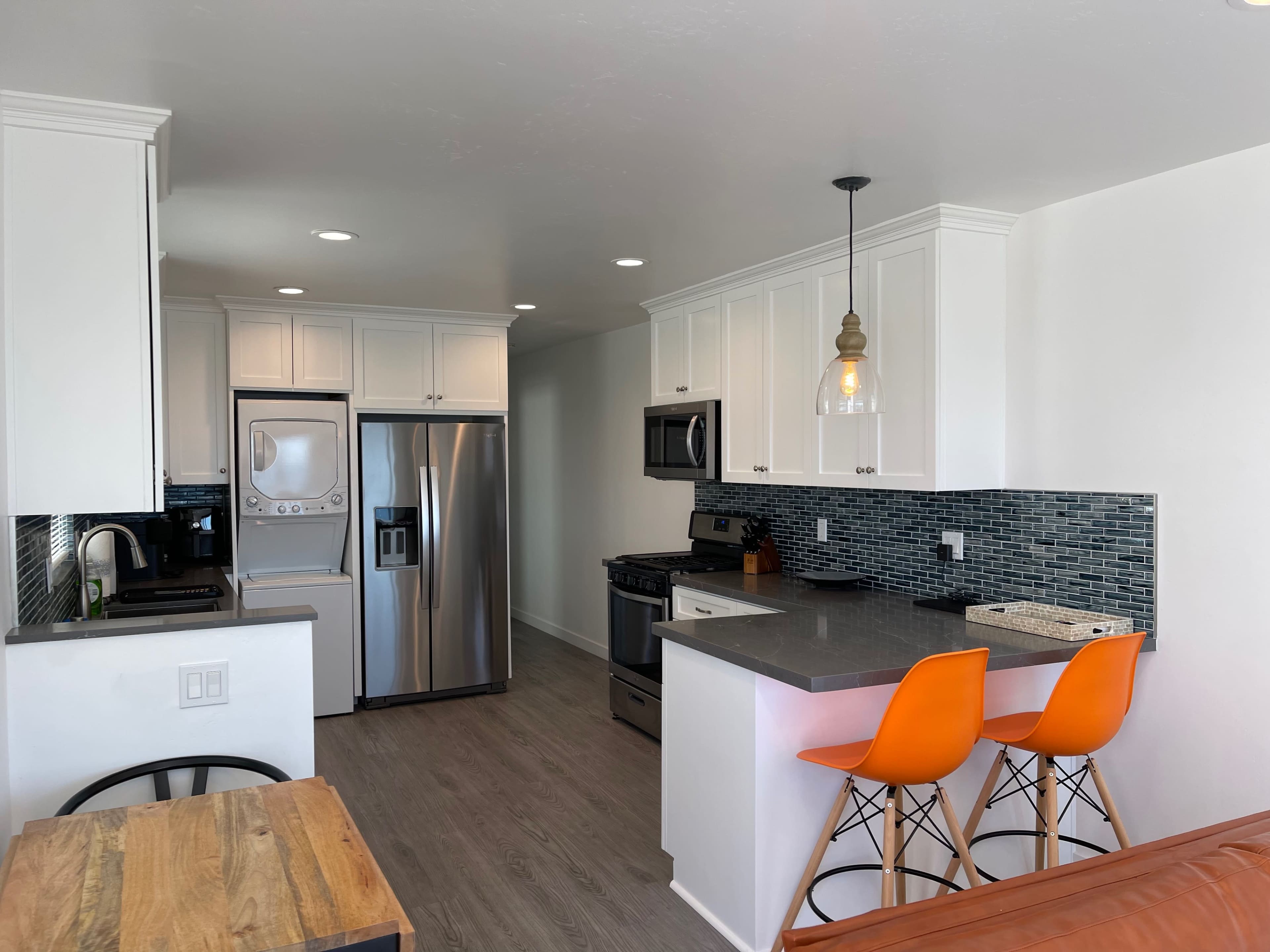 The image shows a modern kitchen with stainless steel appliances, white cabinetry, and a small dining area featuring wooden and orange bar stools.