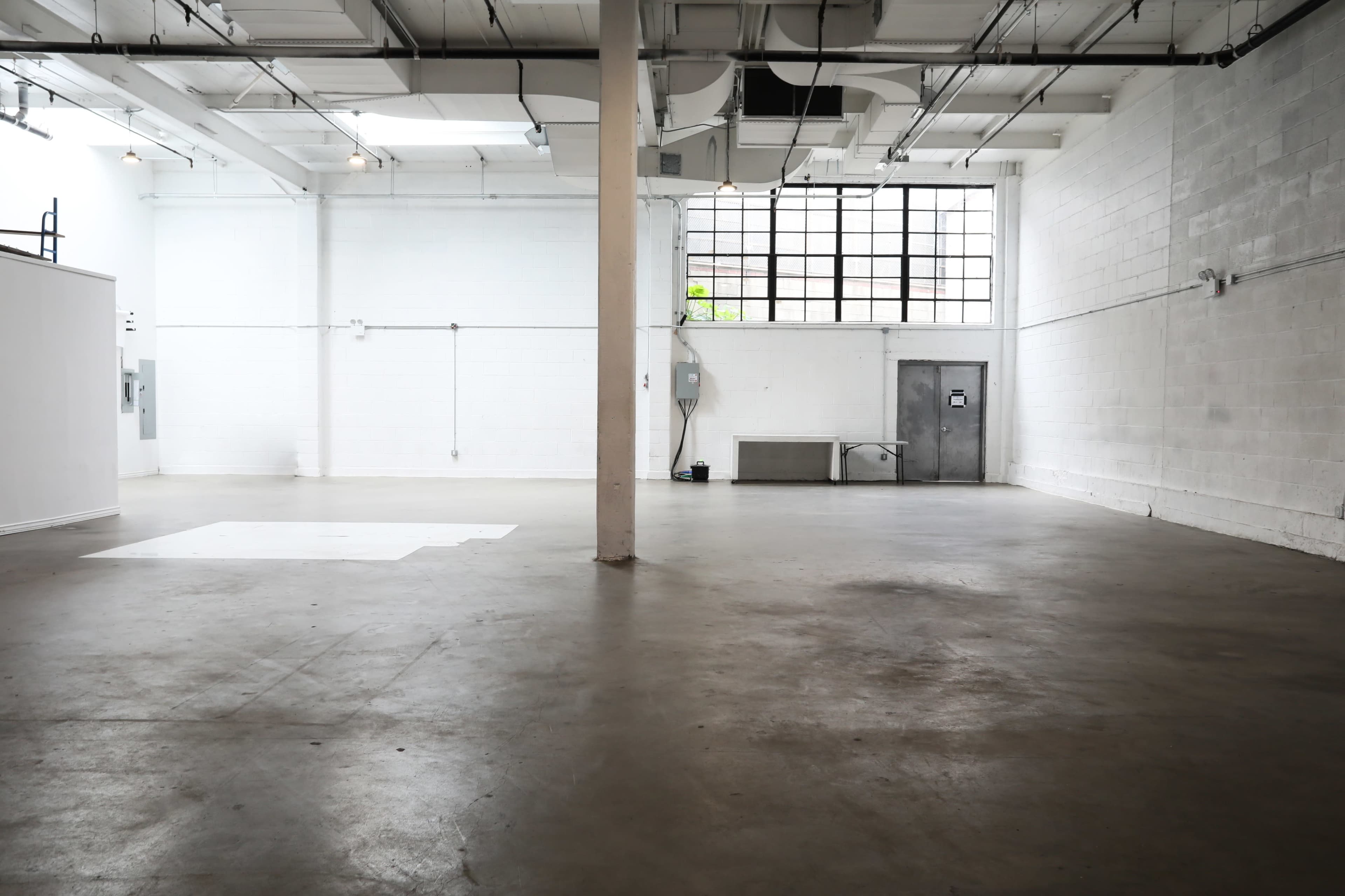 A spacious, empty interior of a warehouse or studio with concrete flooring and a large window on one wall.