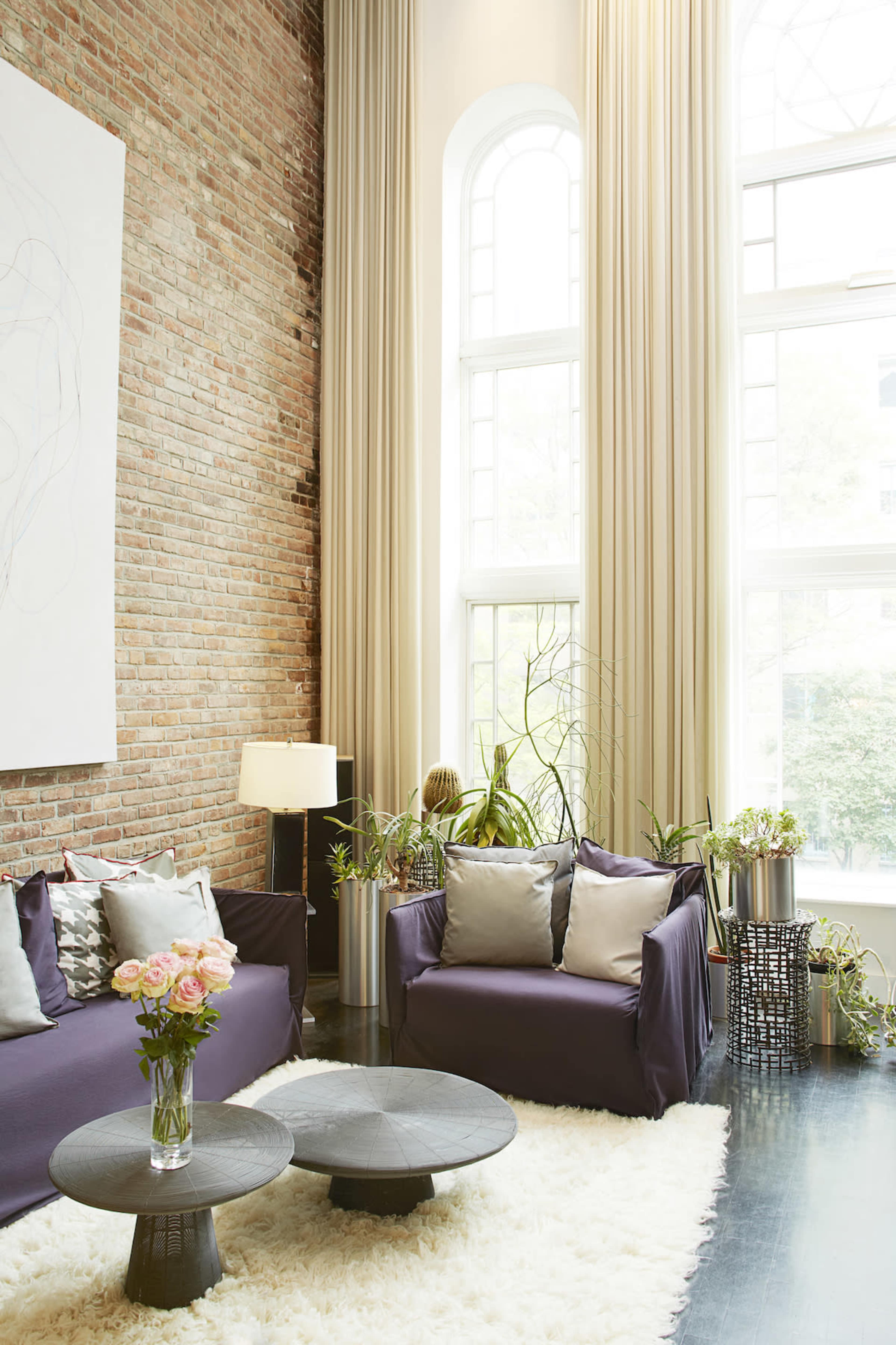 A modern living room features a brick wall, large windows with natural light, two couches, a circular coffee table, and various plants.