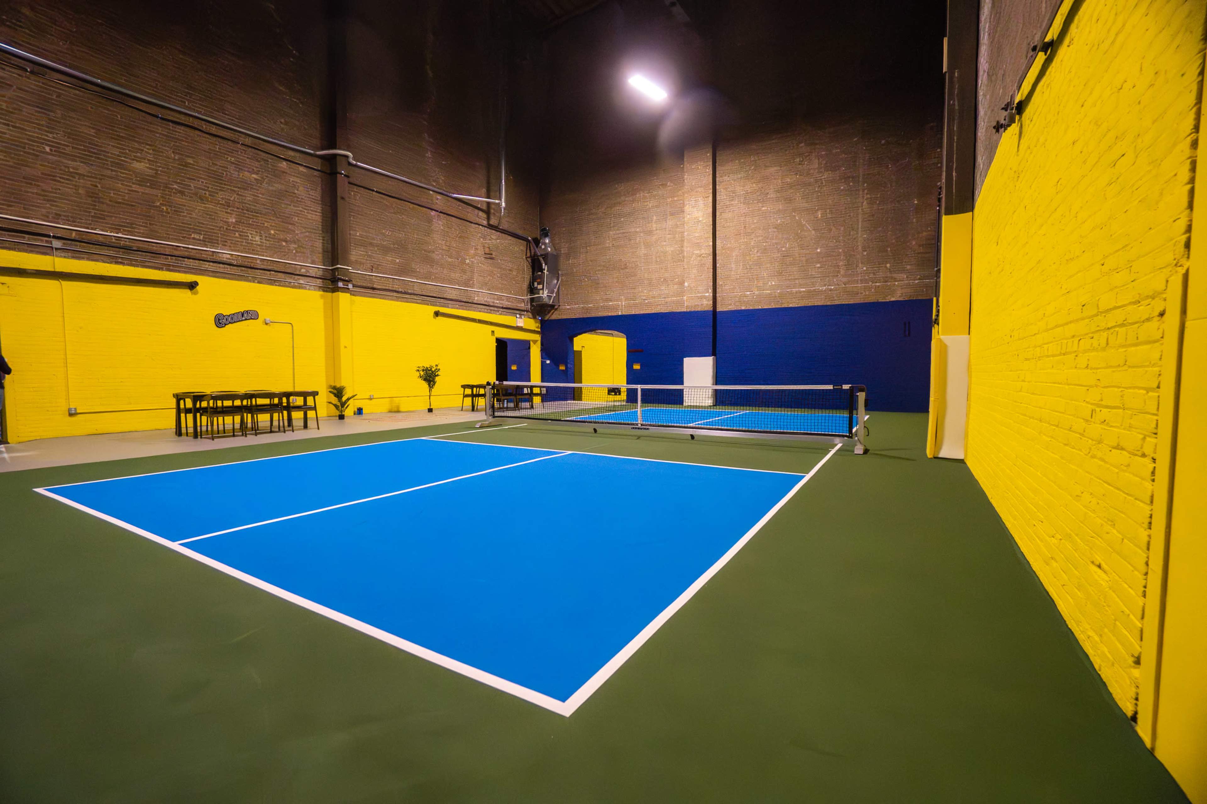 The image shows an indoor tennis court with blue lines on a green surface, surrounded by yellow and blue brick walls.