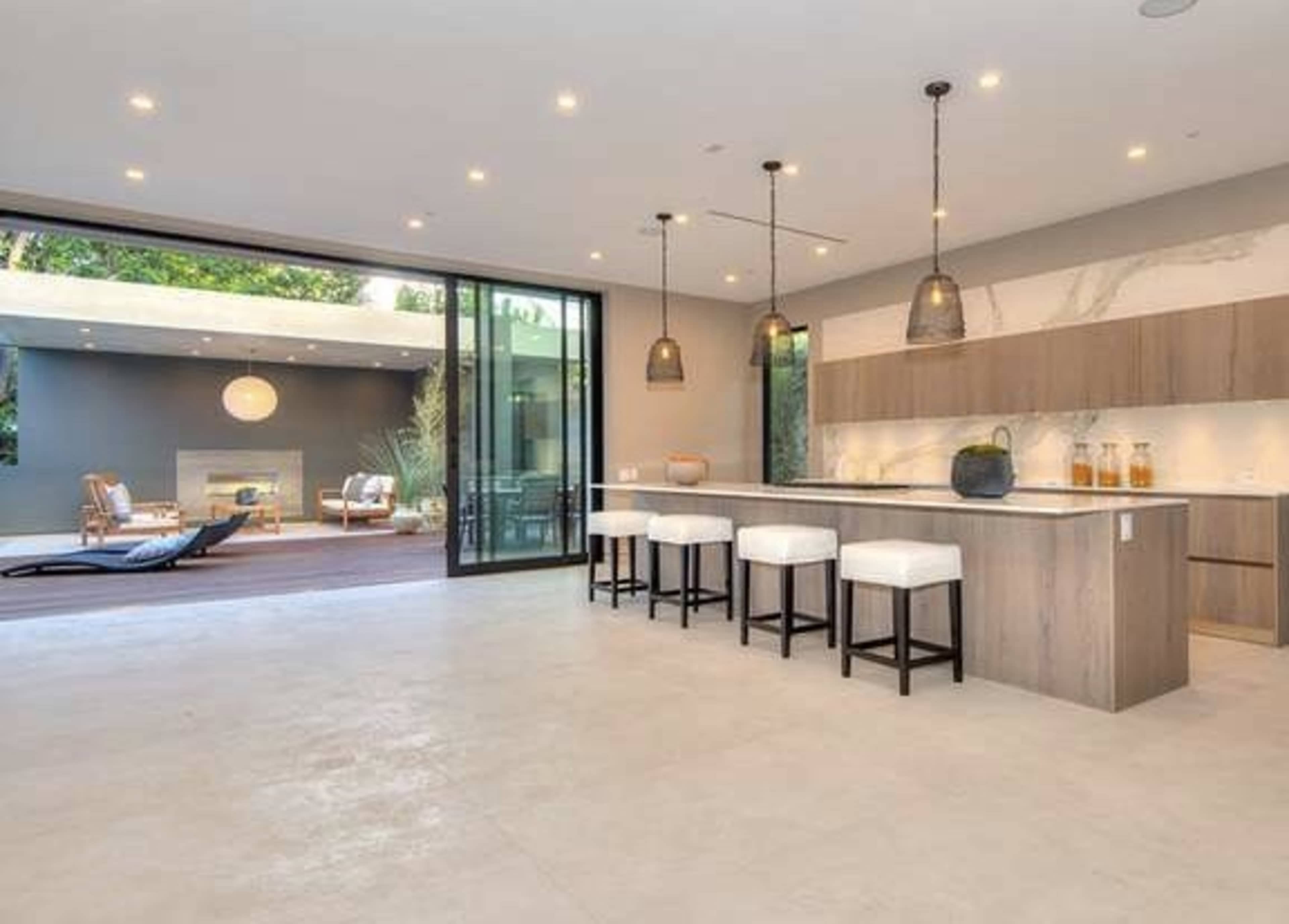 The image shows a modern kitchen area featuring a large island with bar stools, open to a living space with a patio in the background.