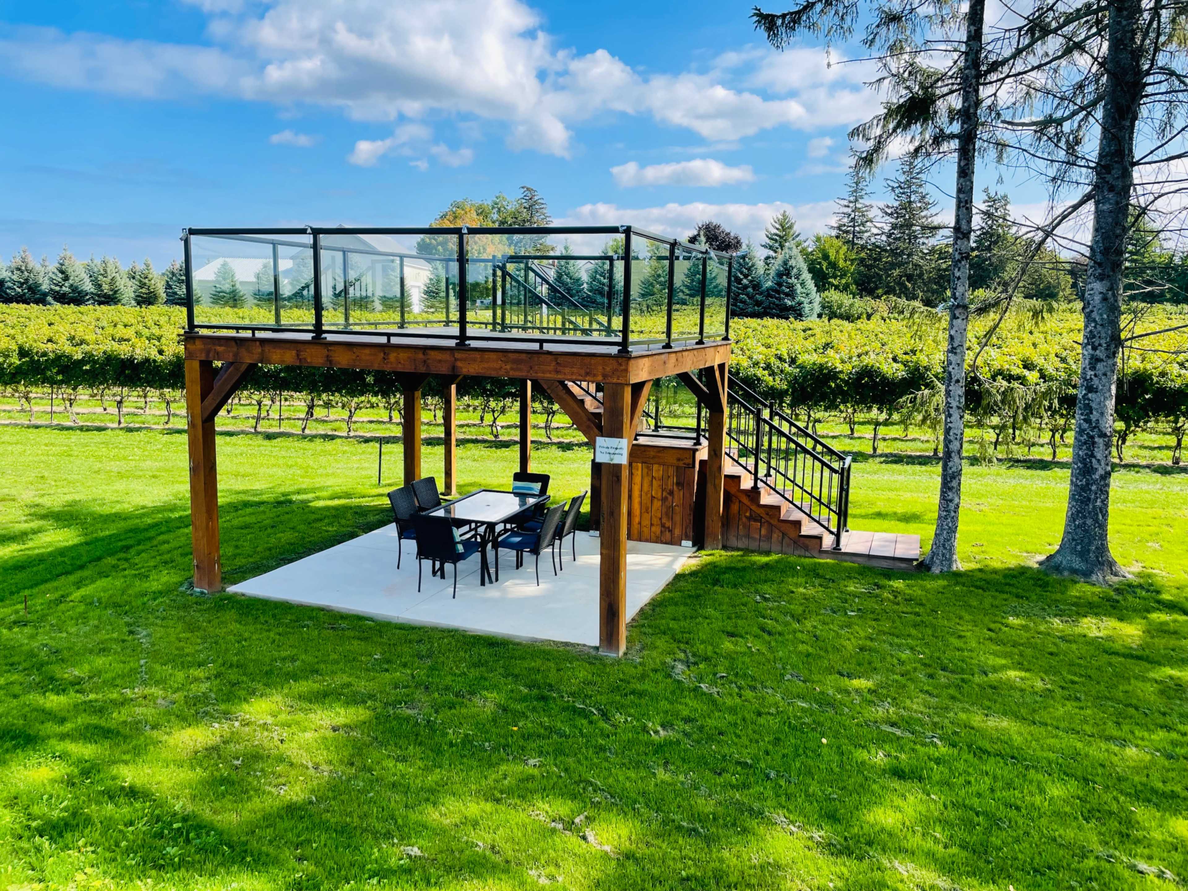 A wooden deck with a glass railing is elevated above a patio area featuring a dining set, surrounded by a grassy lawn and vineyards.