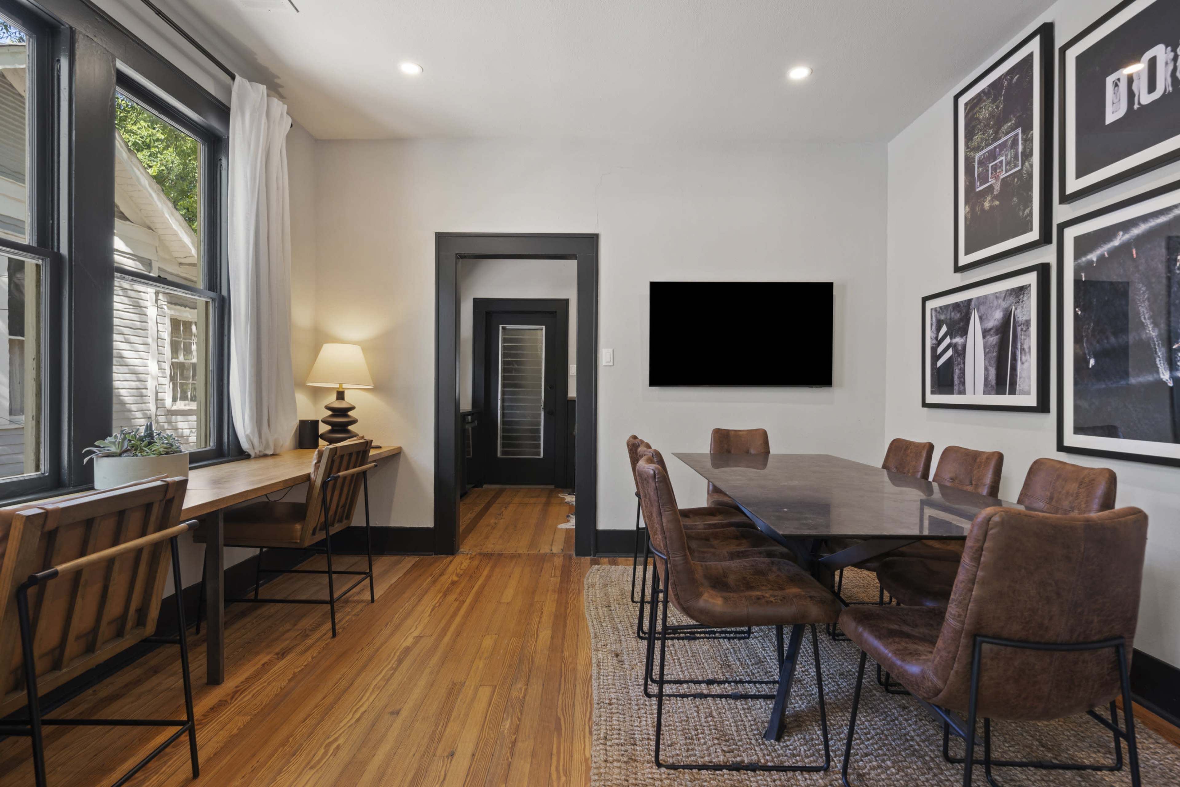 A dining area features a long table surrounded by brown chairs, with large windows and a wall adorned with framed photographs.