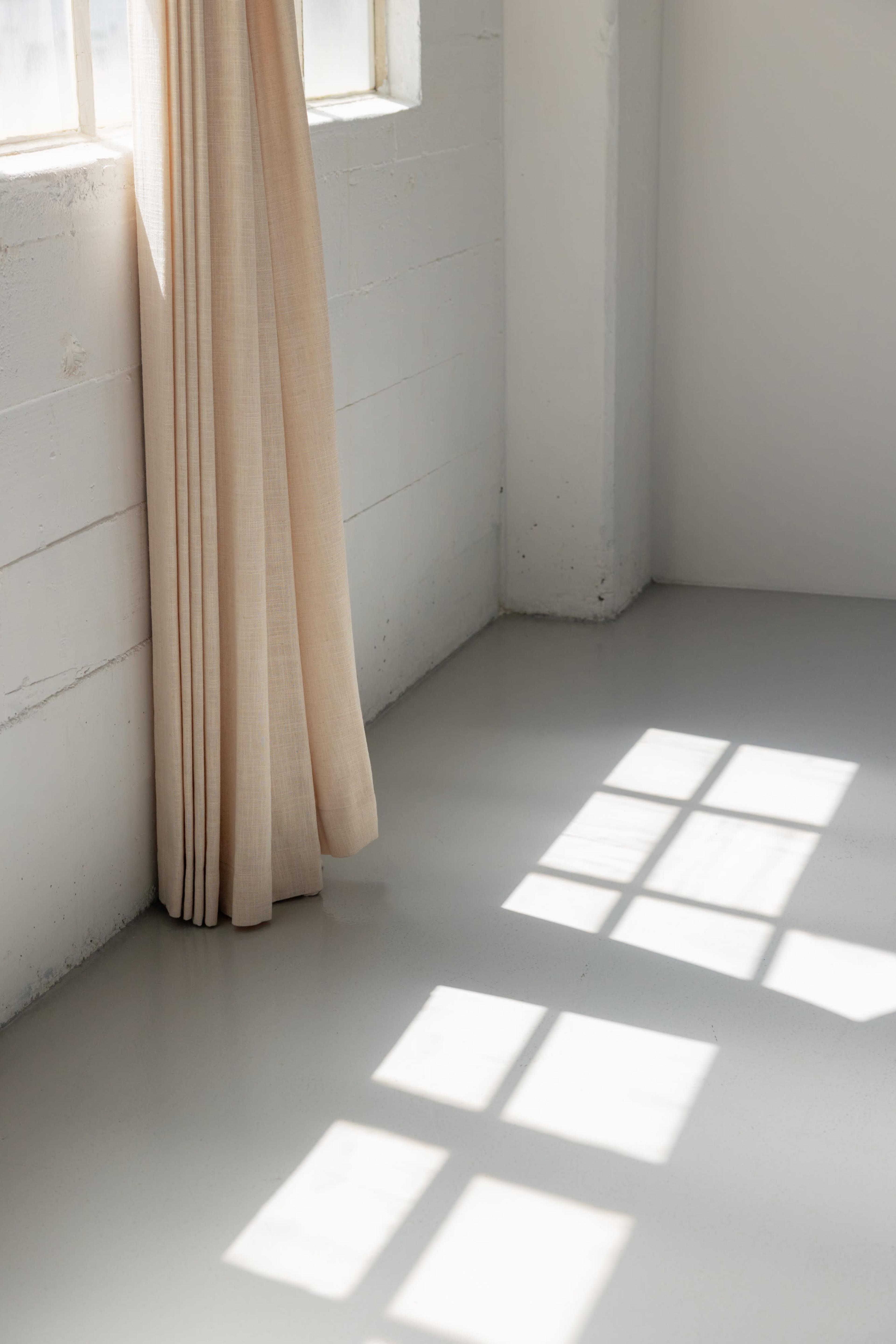 The image shows a corner of a room with light-colored curtains hanging beside a window, casting square shadows on the smooth floor.