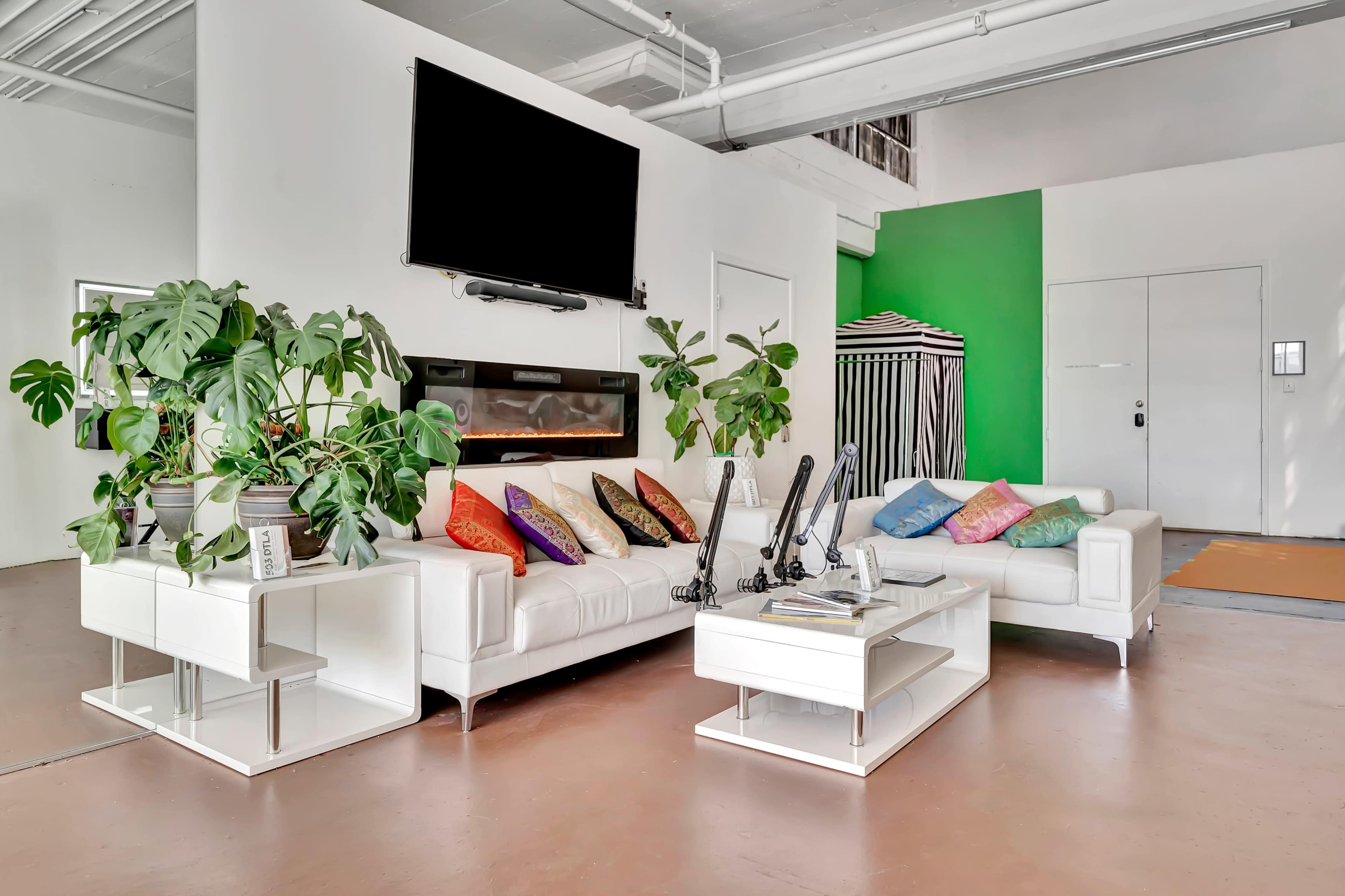 A modern living room features a white sectional sofa with colorful cushions, a flat-screen TV mounted on a white wall, and a green accent wall behind a standing striped umbrella.