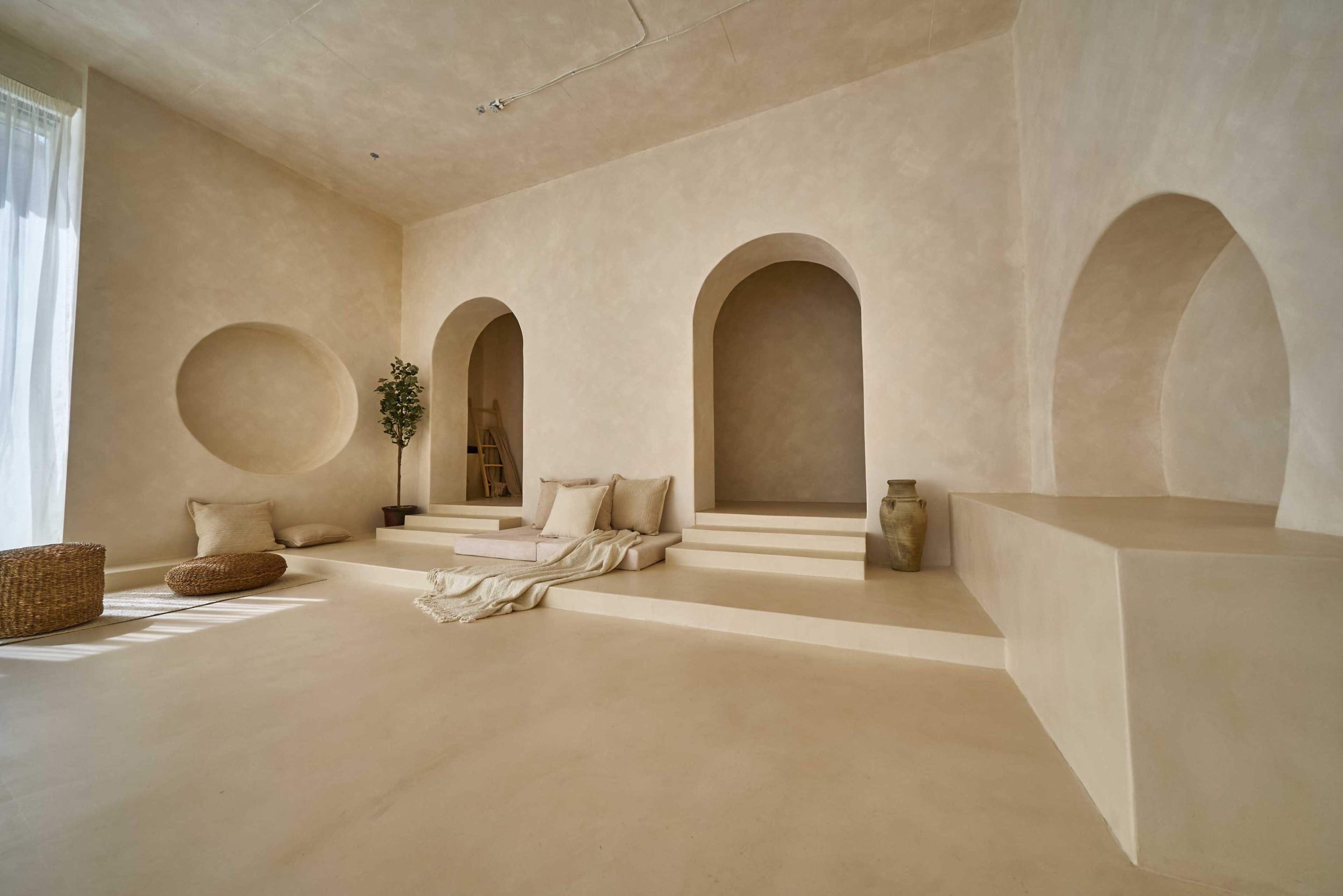 The image features a minimalist interior space with textured beige walls, arched openings, a staircase, and simple furniture elements including cushions and a woven basket.