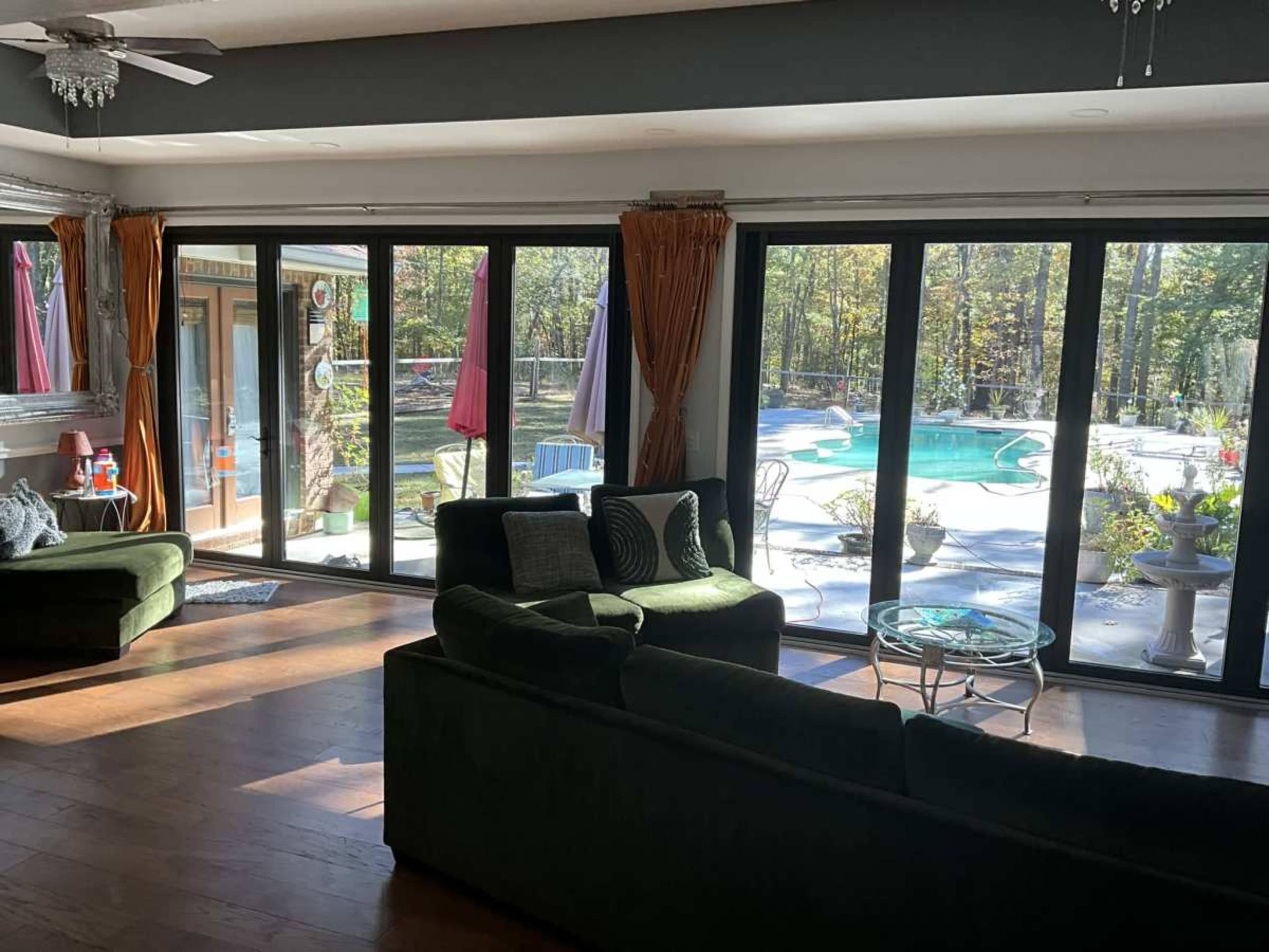 The interior of a room features green sofas and large windows overlooking a backyard with a swimming pool surrounded by trees.