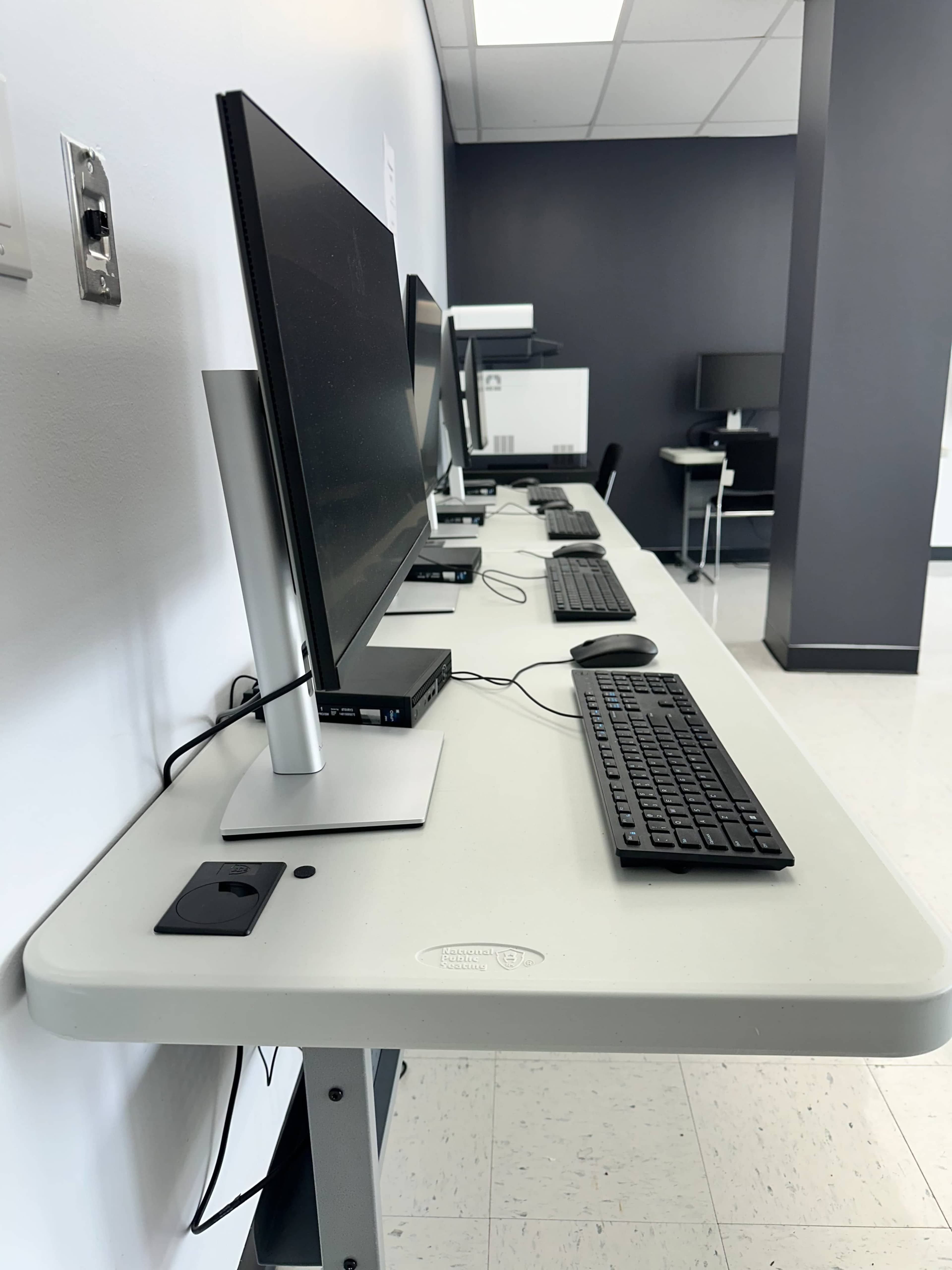 A row of desktop computers with monitors and keyboards is set up on a long white table in a well-lit room.
