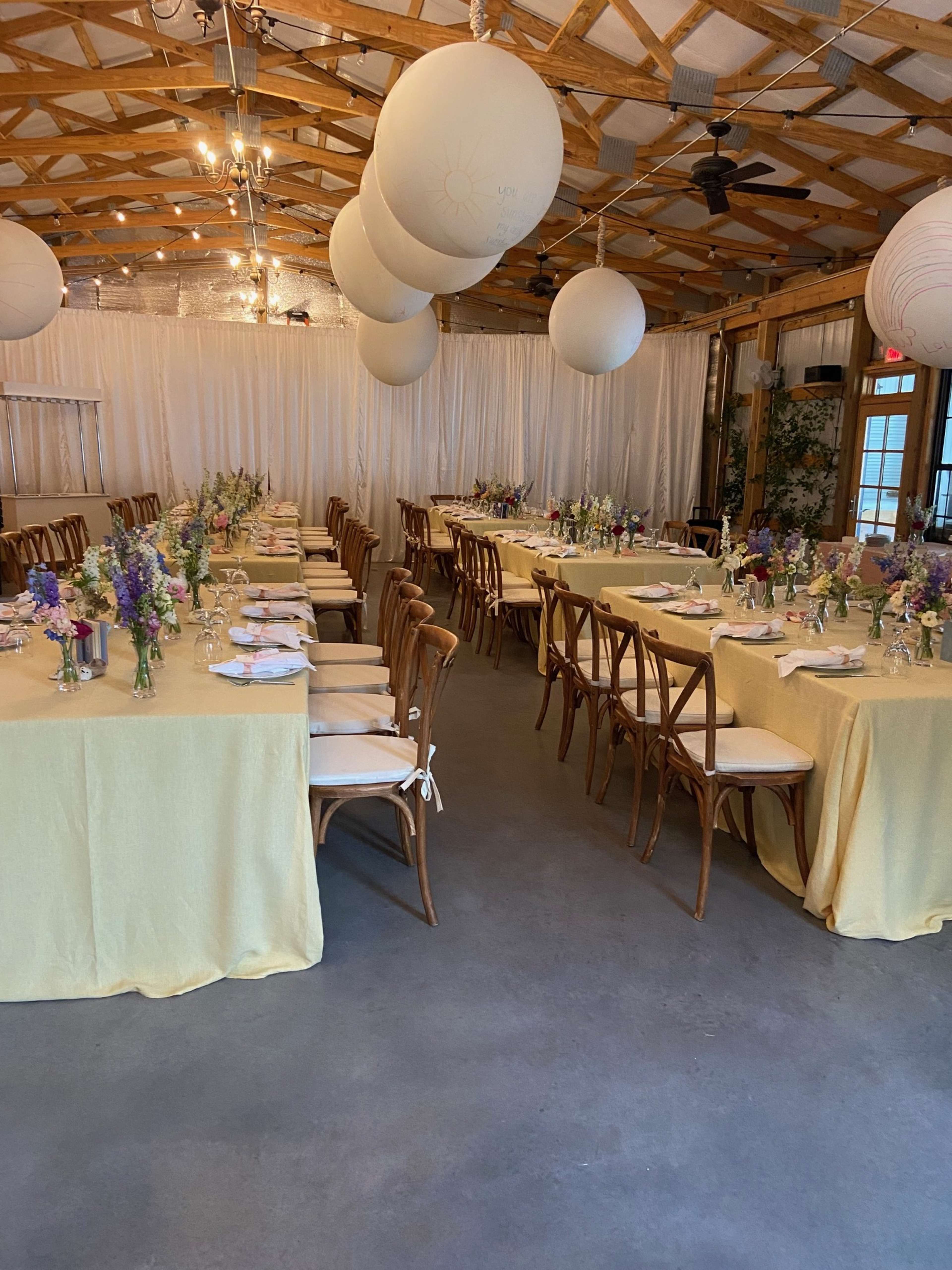 The image shows a reception area with long tables covered in yellow tablecloths, arranged with flower centerpieces, and decorated with large white paper lanterns overhead.
