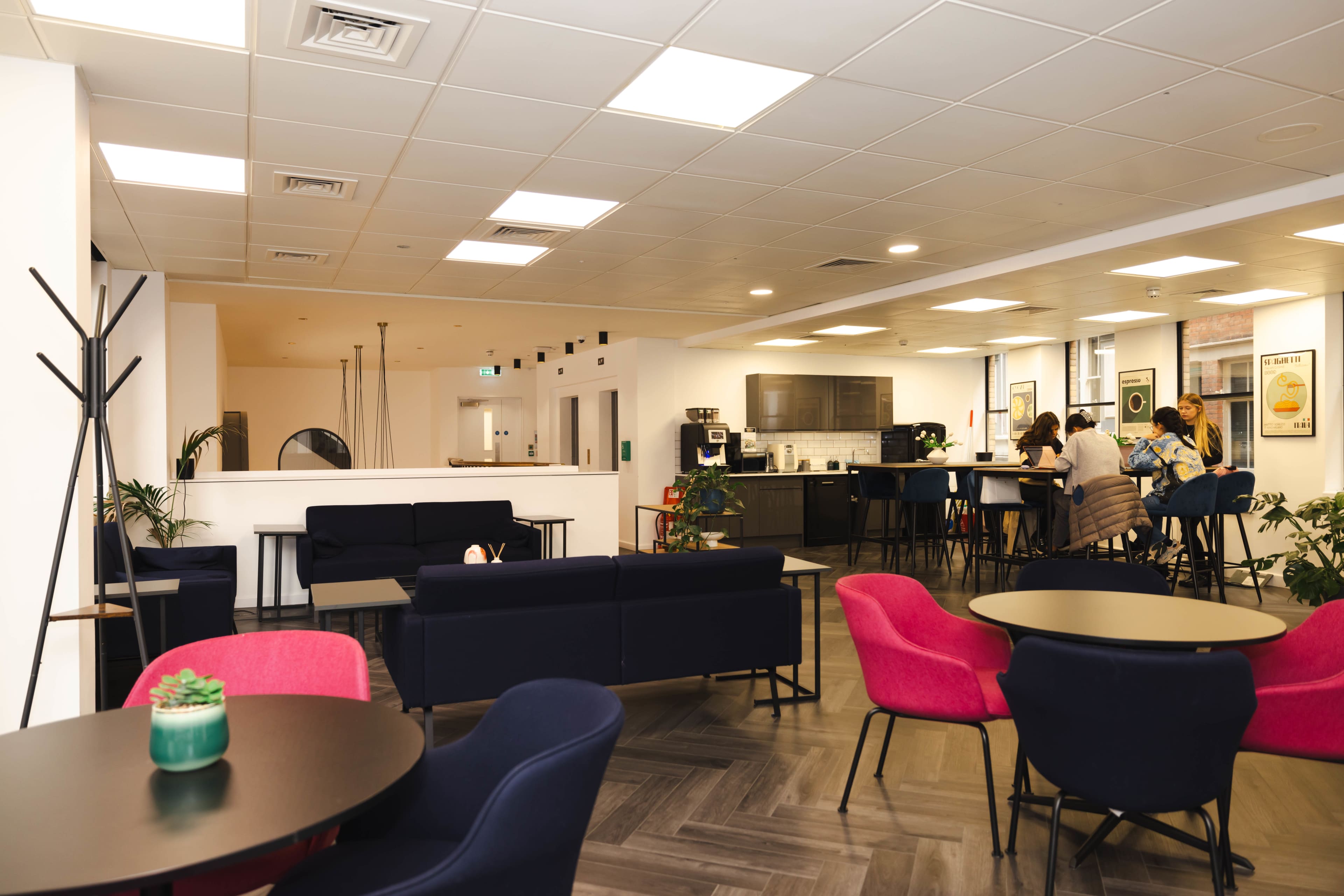 A modern lounge area with seating in various colors, a kitchenette, and several people discussing at a tall table.