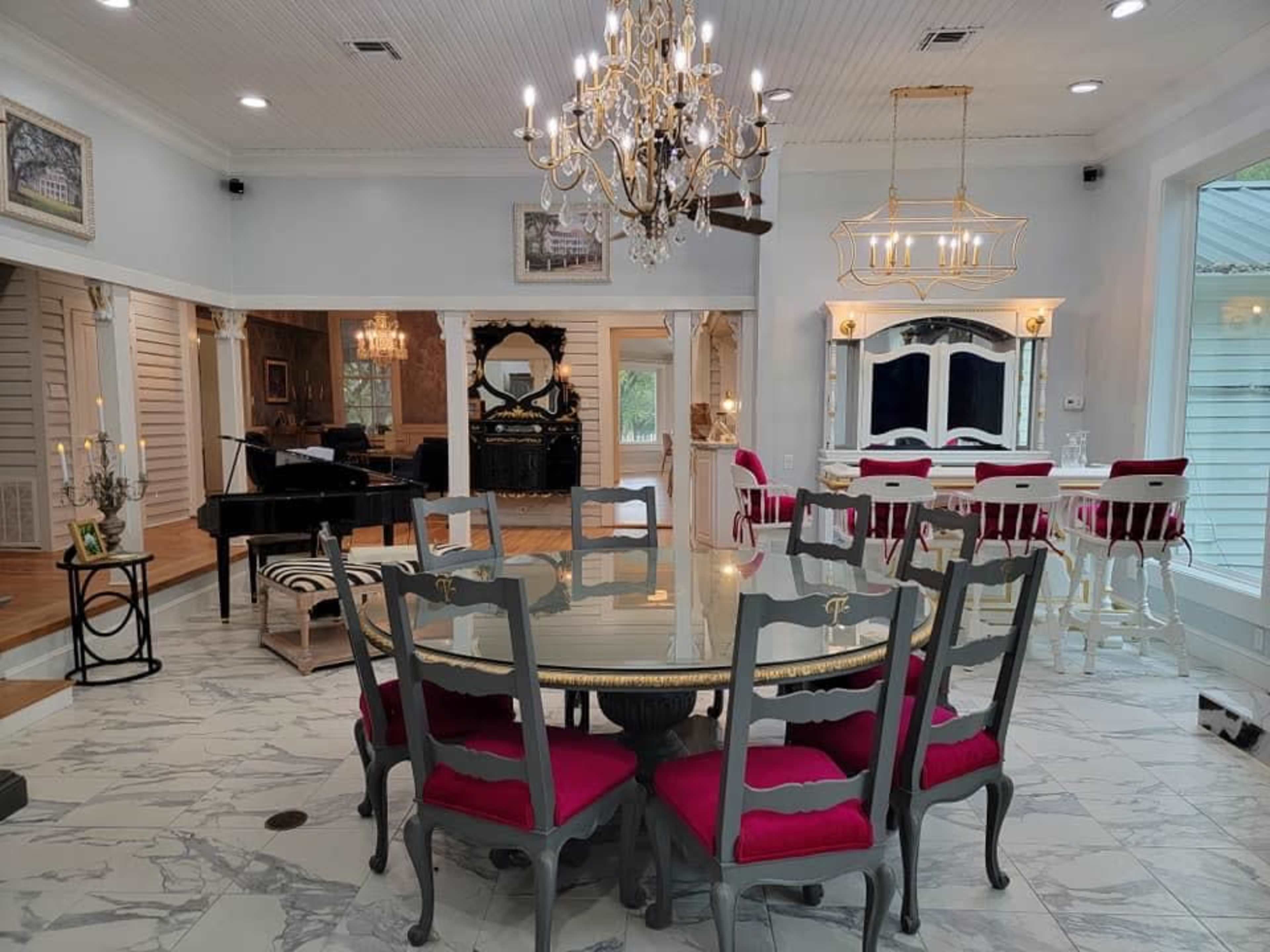 A spacious dining area featuring a round table with gray chairs and red cushions, a chandelier overhead, and a view of a grand piano in an adjacent room.