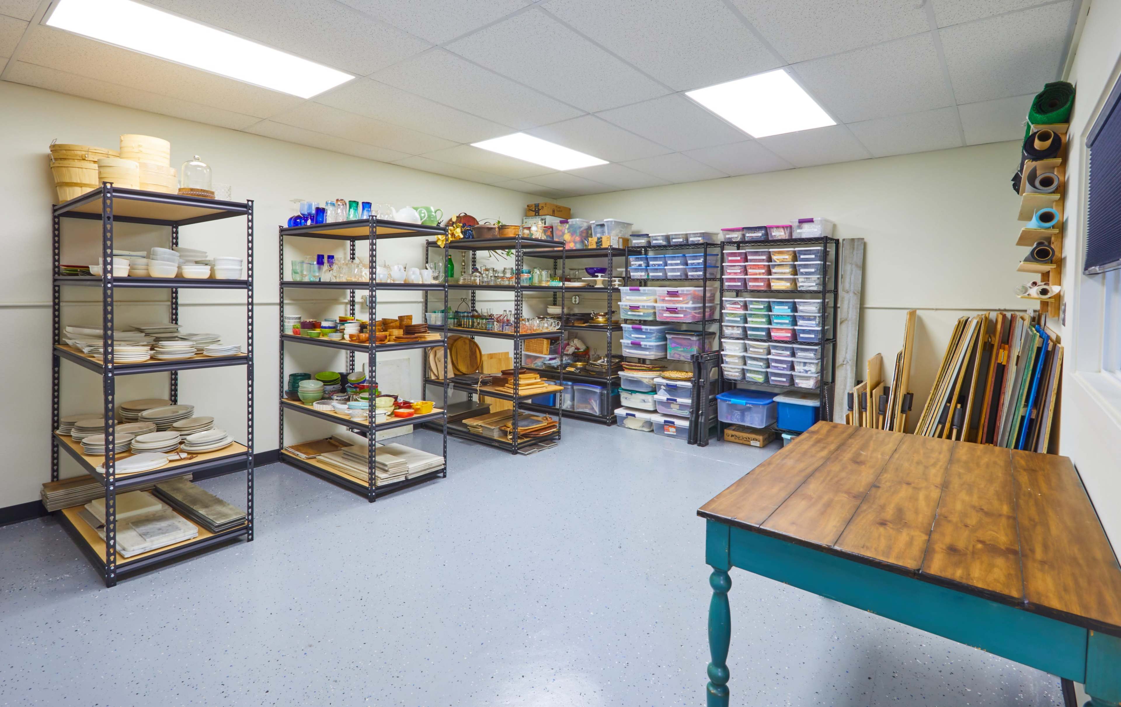 A well-organized storage room features shelving units filled with various containers, dishes, and utensils, along with a wooden table and several rolled items along the walls.