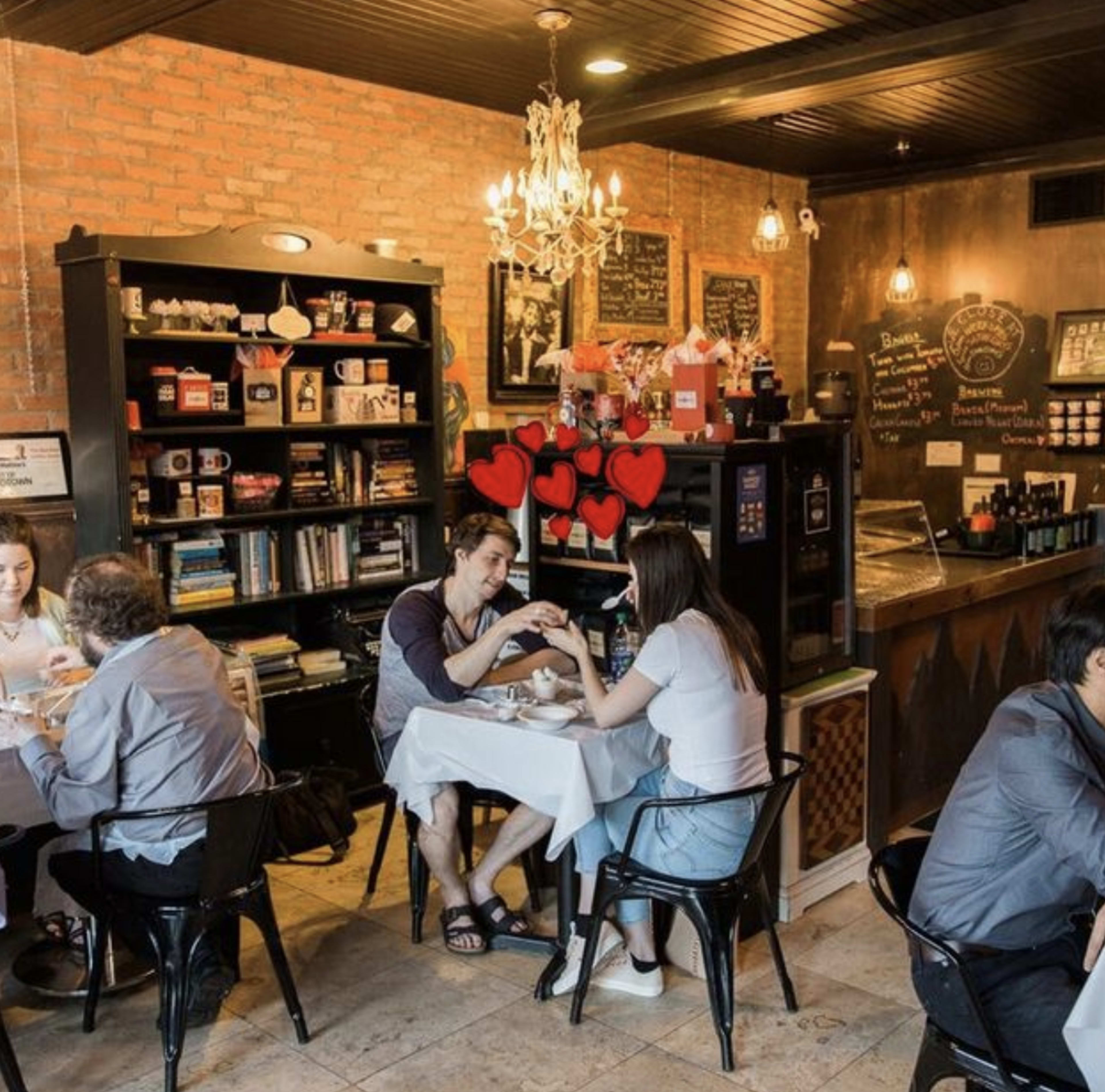 A couple sits at a small table in a cozy café, surrounded by shelves filled with various items, while other patrons enjoy their meals nearby.