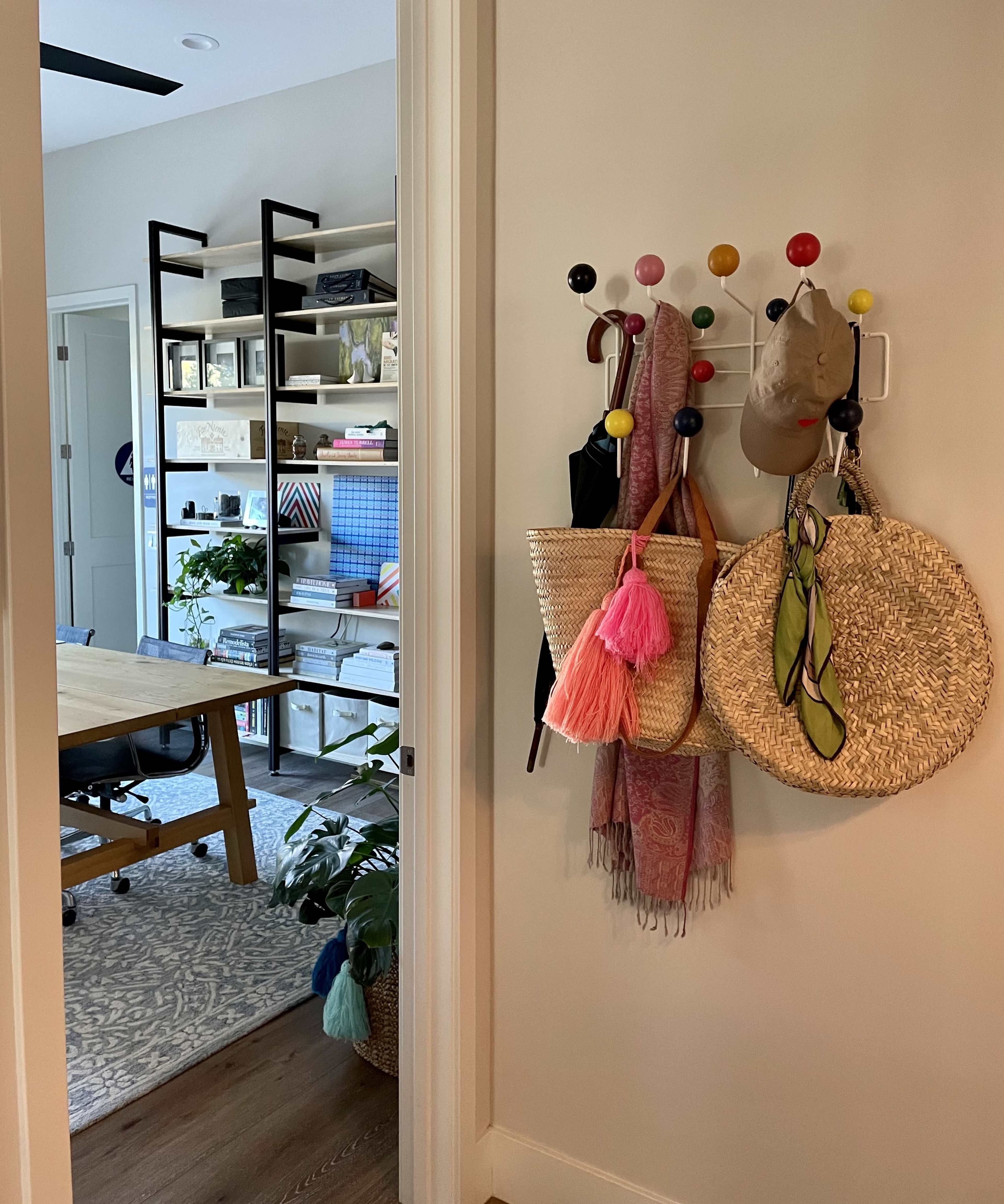 The image shows a hallway with a coat rack featuring bags and a hat, leading to a room with a desk and shelves filled with books and plants.