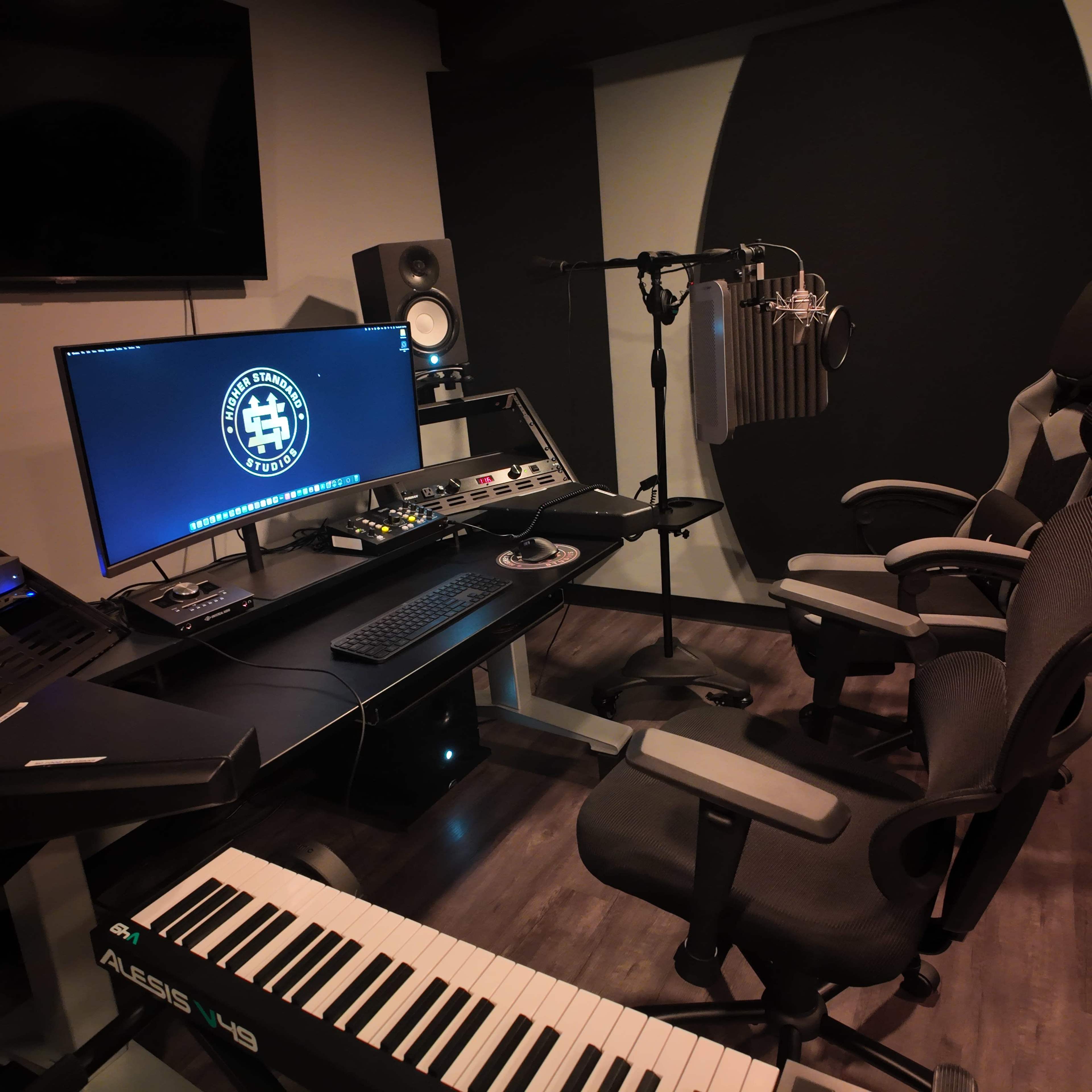 The image shows a modern recording studio with a digital audio workstation setup, a microphone on a boom arm, and an Alesis keyboard in the foreground.