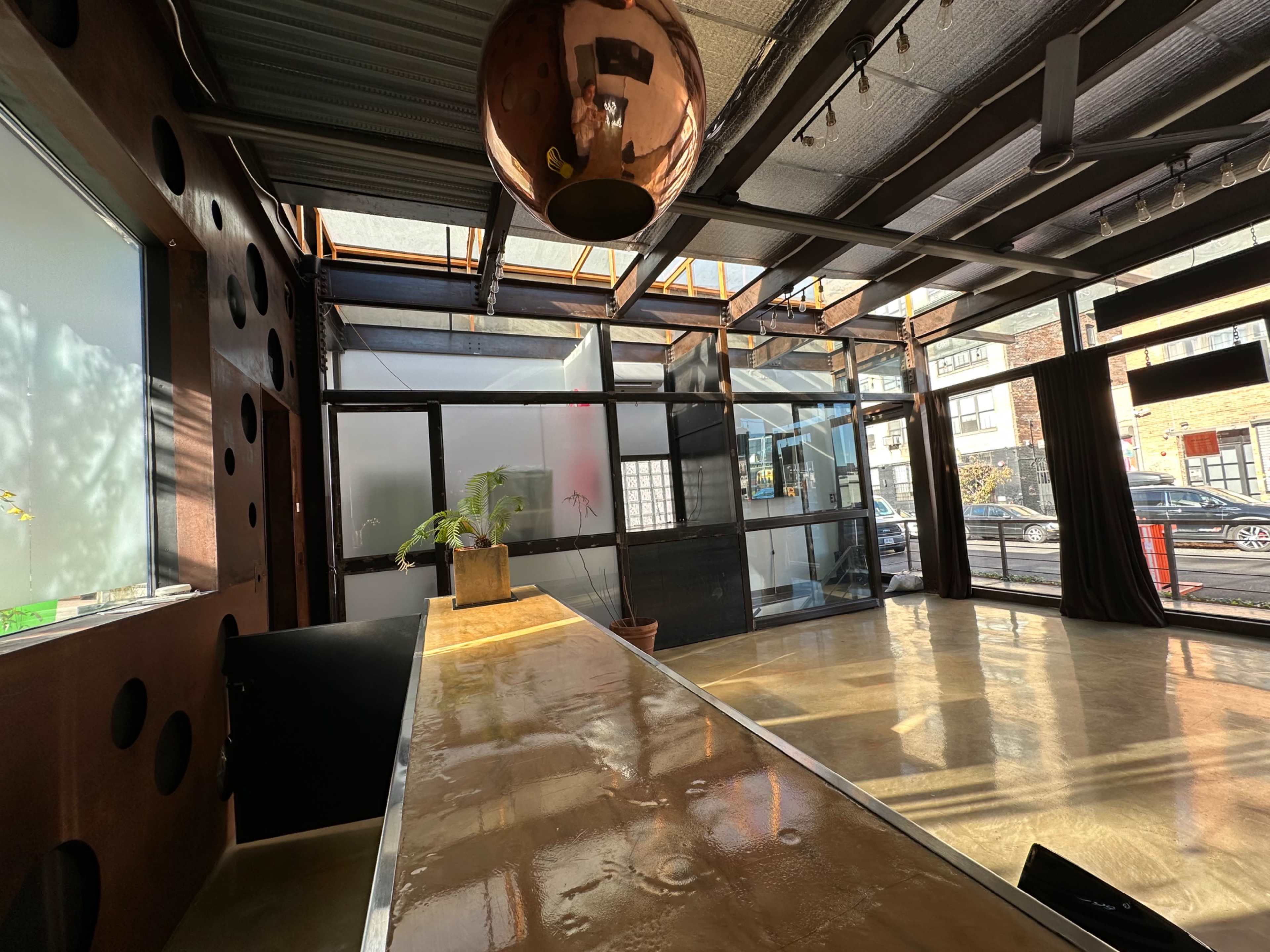 The image shows a modern interior space with a long, polished counter, large windows, and a minimalistic design featuring a potted plant and a reflective copper light fixture.