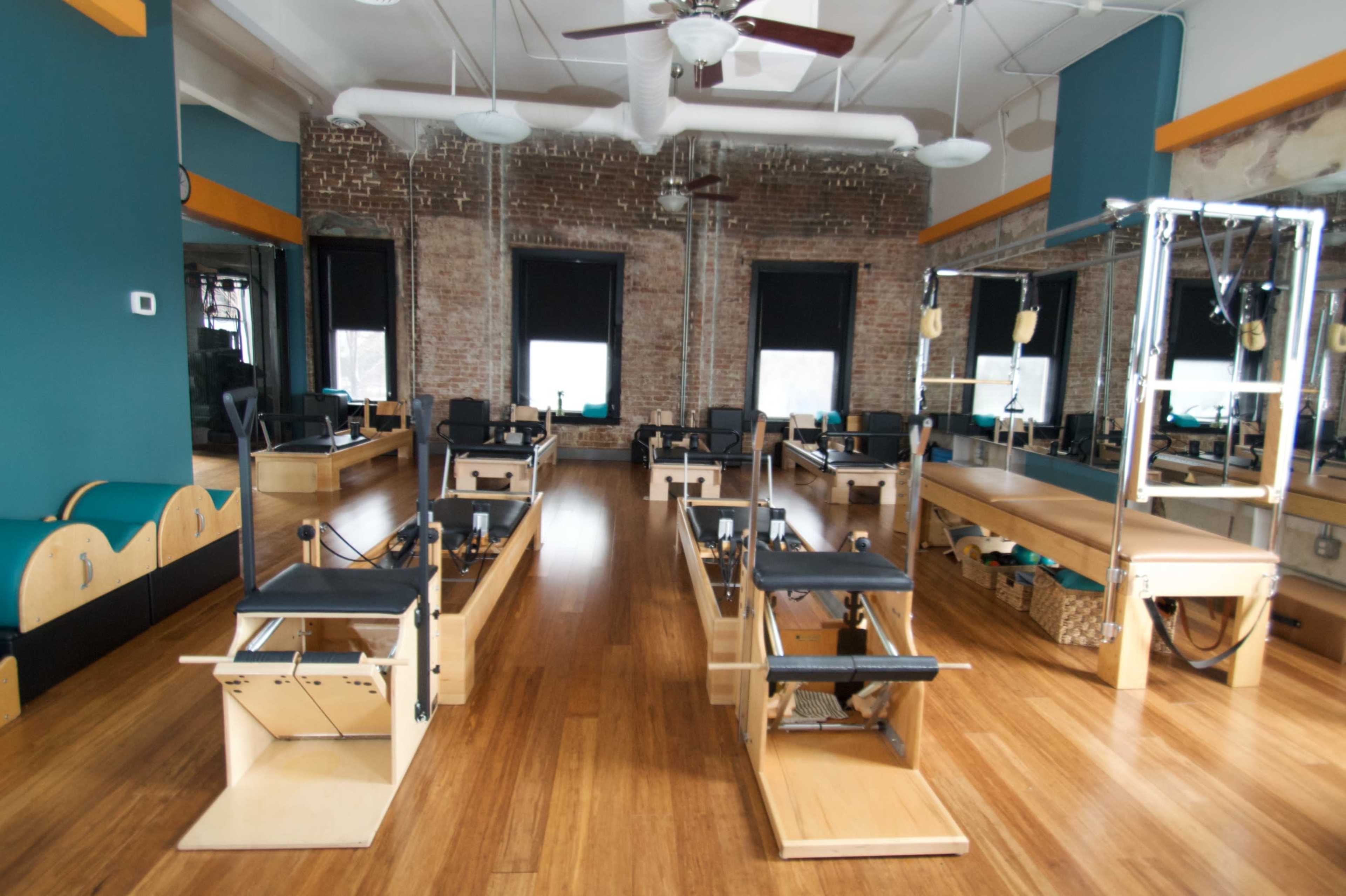 A spacious Pilates studio with wooden floors, several reformer machines, and brick walls.