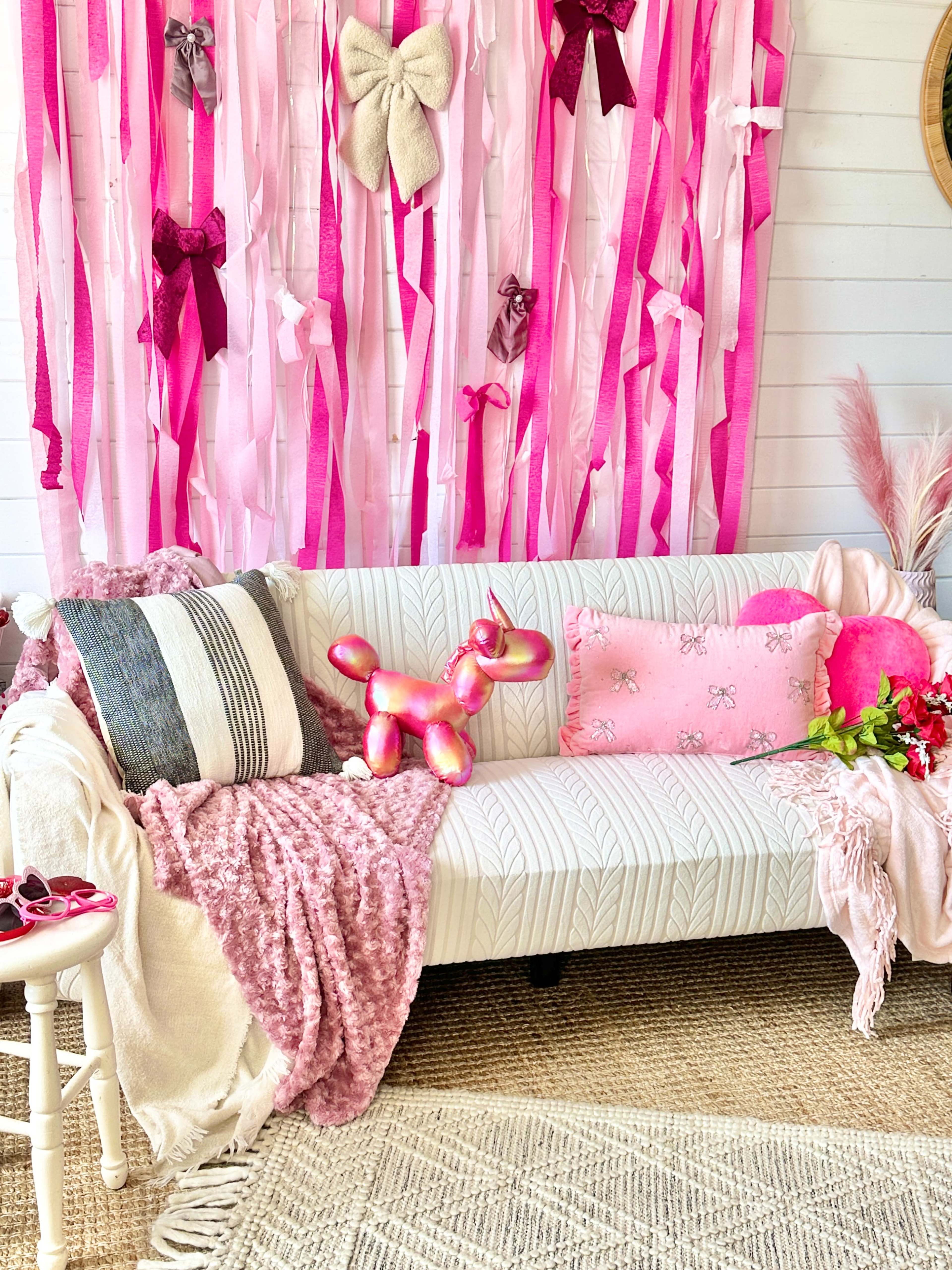 The image shows a white couch adorned with various pink and purple decorations, including pillows, a throw blanket, and a backdrop of pink ribbons.