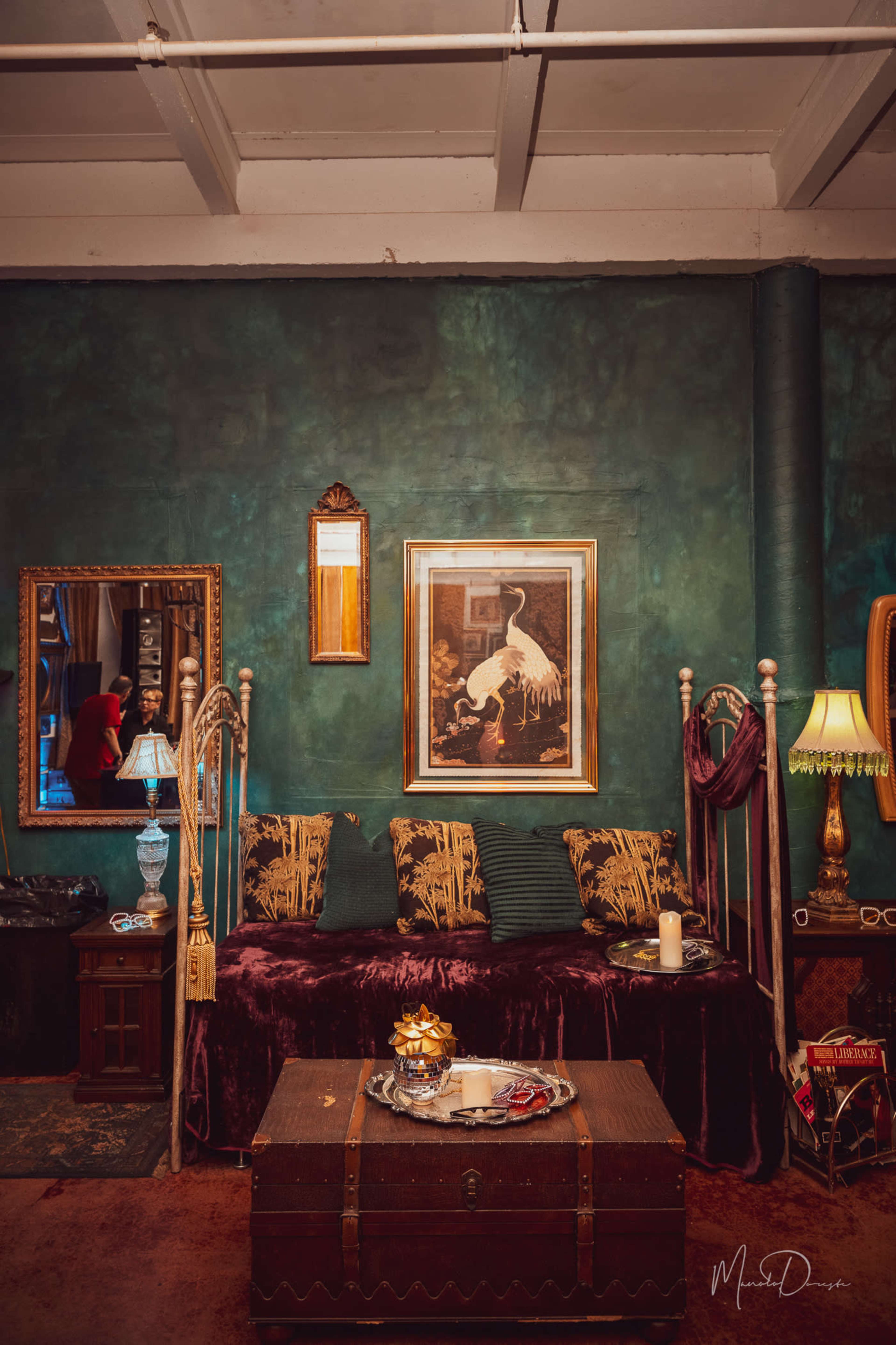 A cozy room with a green wall, a decorative sofa adorned with cushions, a wooden trunk in front, and a mirror reflecting a person in the background.