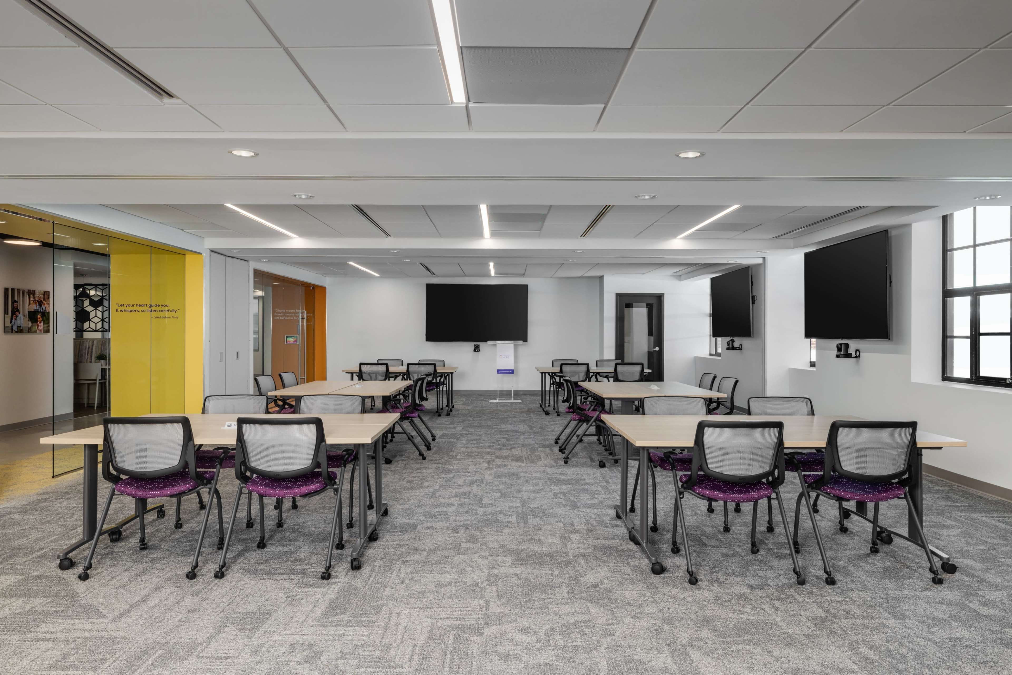 The image shows a modern classroom or meeting room with multiple tables arranged in a grid layout and several chairs, along with wall-mounted screens.