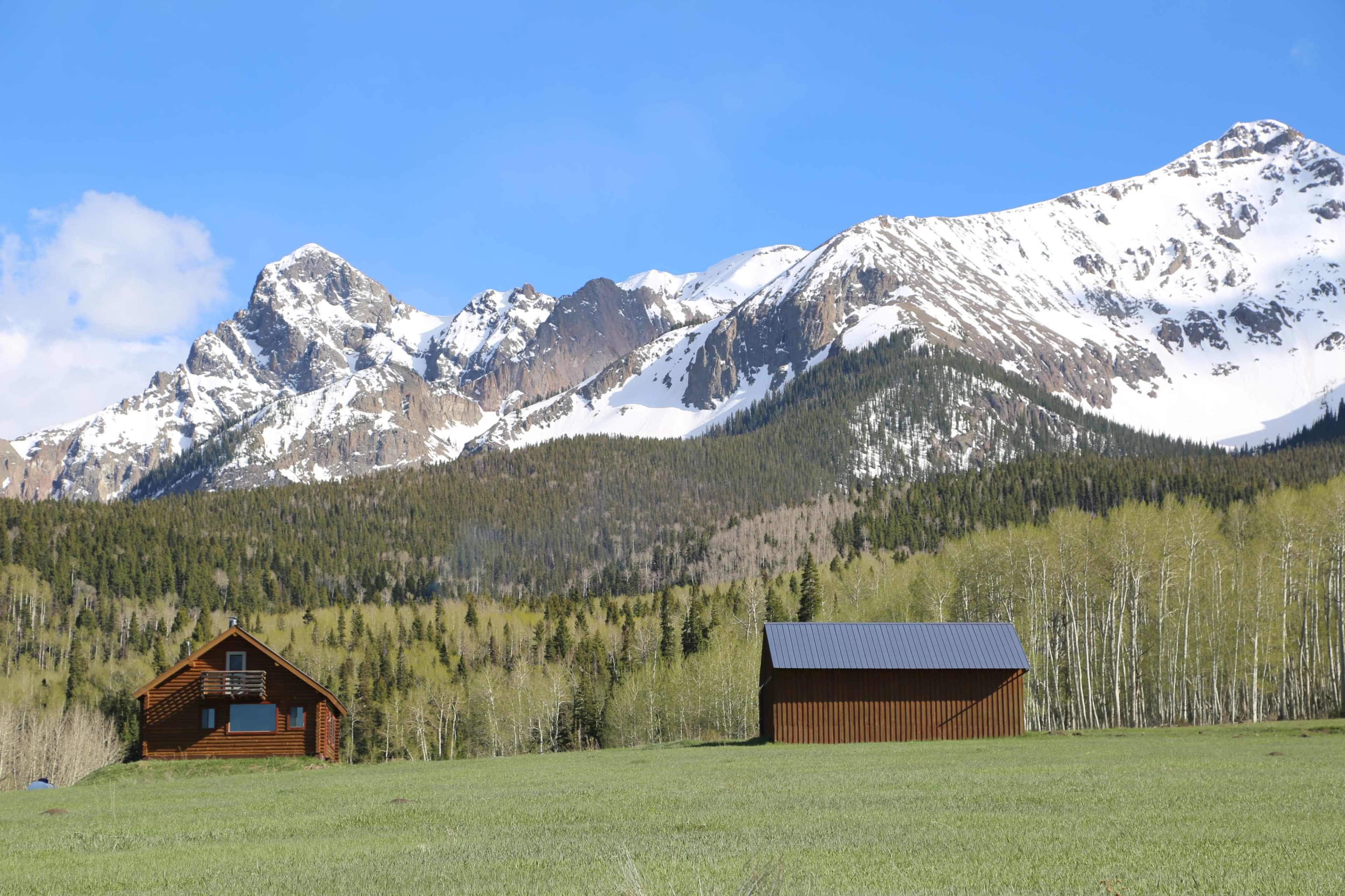 Two wooden cabins sit in a green meadow with snow-capped mountains in the background.