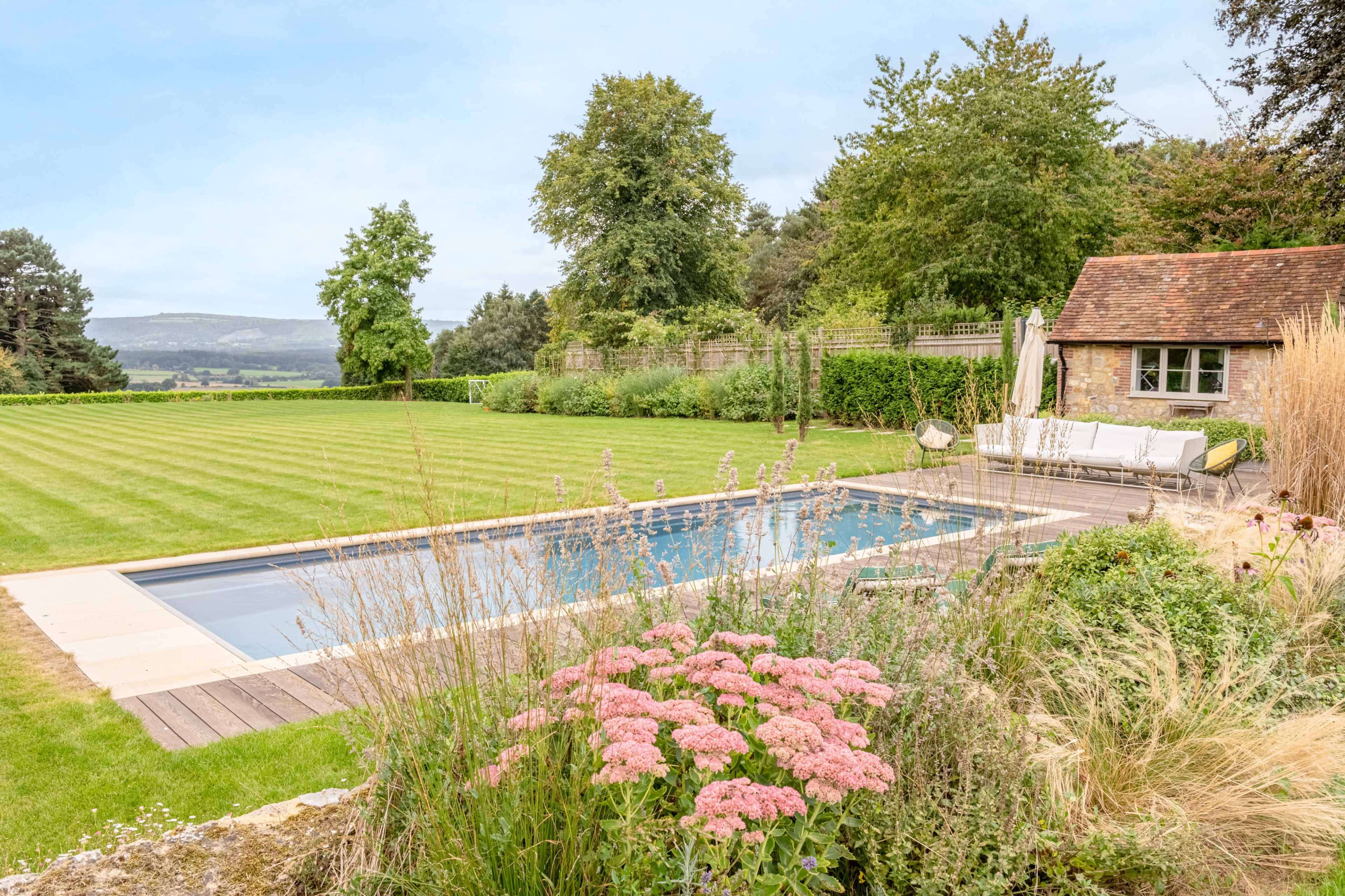 A rectangular swimming pool with lounge chairs is surrounded by neatly trimmed grass and flower beds in a landscaped garden.