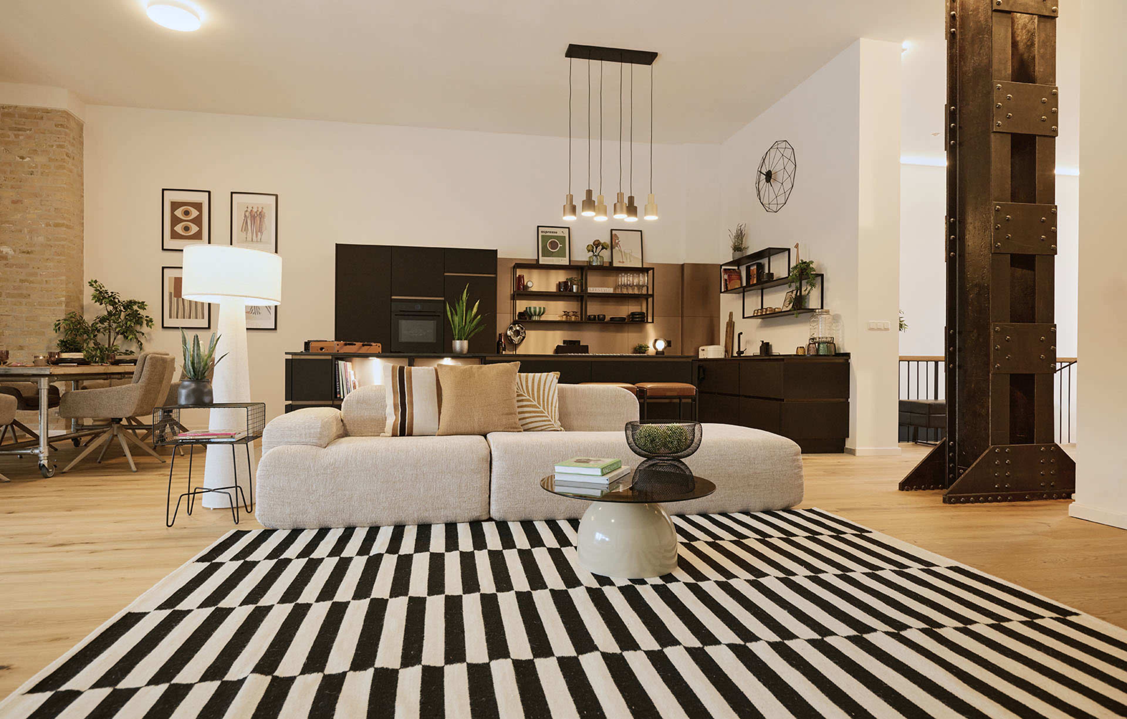 The image shows a modern living room with a sectional sofa on a striped rug, featuring a dining area in the background and minimalist decor elements.