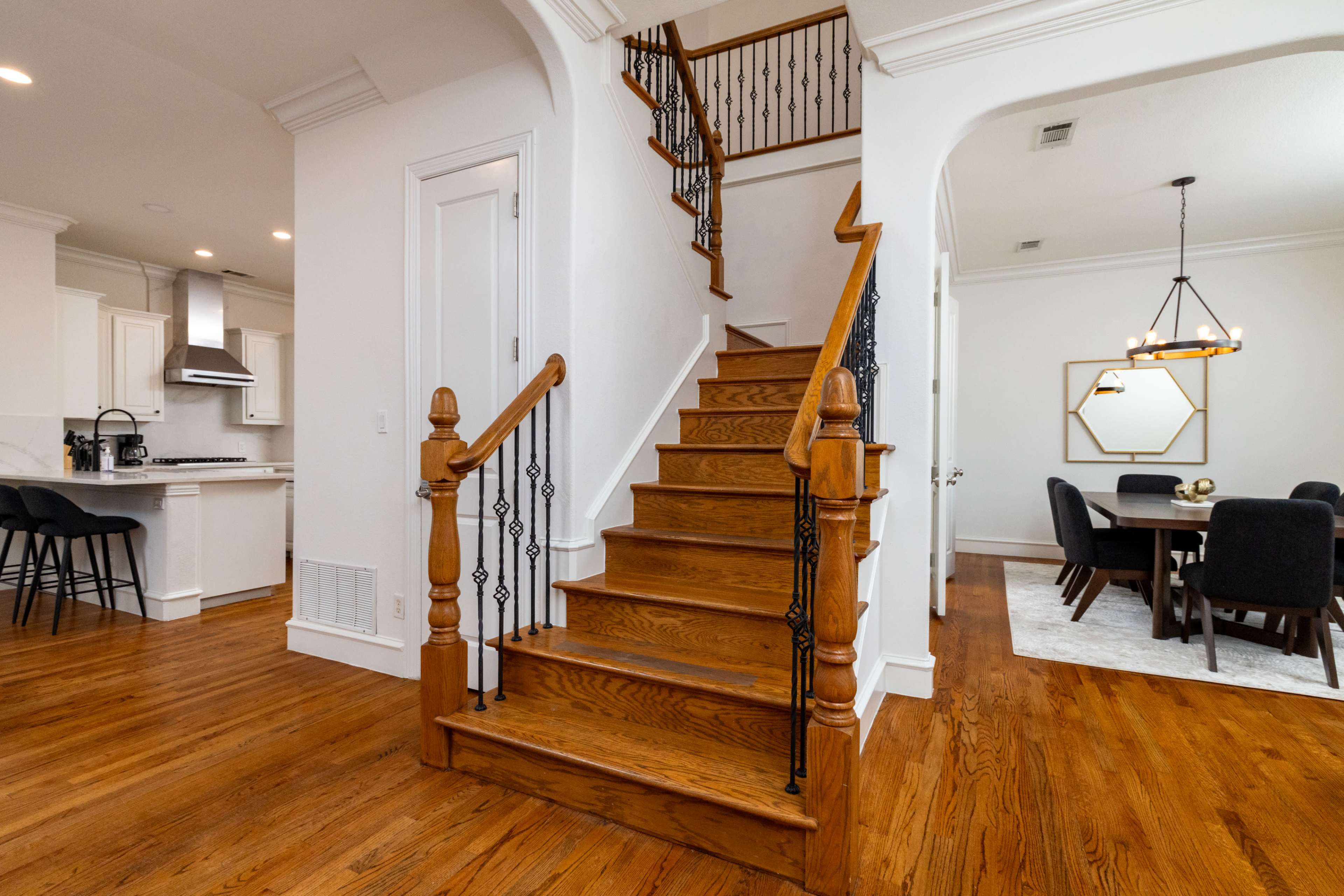 The image shows a wooden staircase with a decorative black wrought iron railing, leading to an upper level, alongside a dining area and a modern kitchen in an open floor plan.