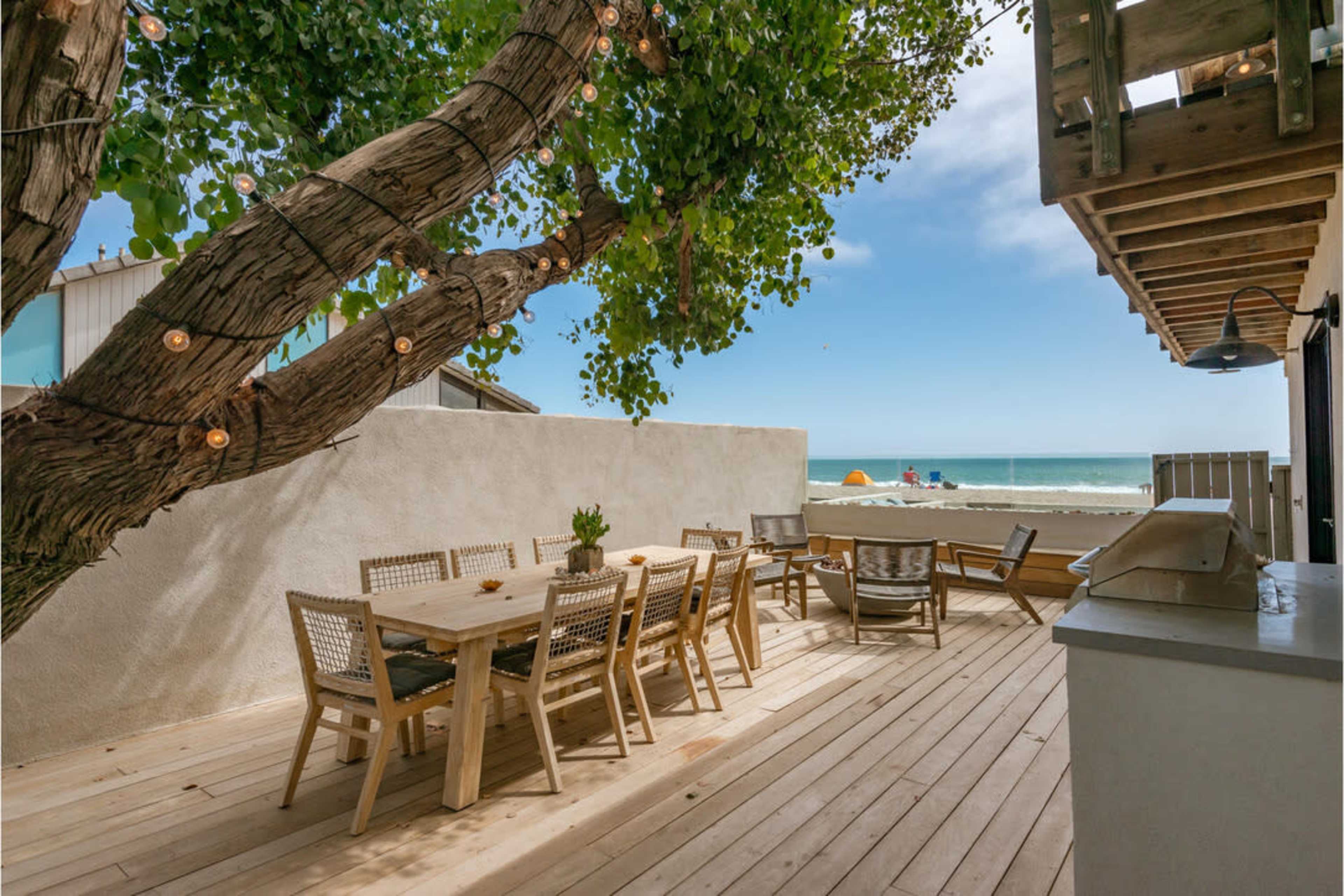A wooden deck with a dining table and chairs overlooks the ocean, with a tree providing shade and string lights hanging above.