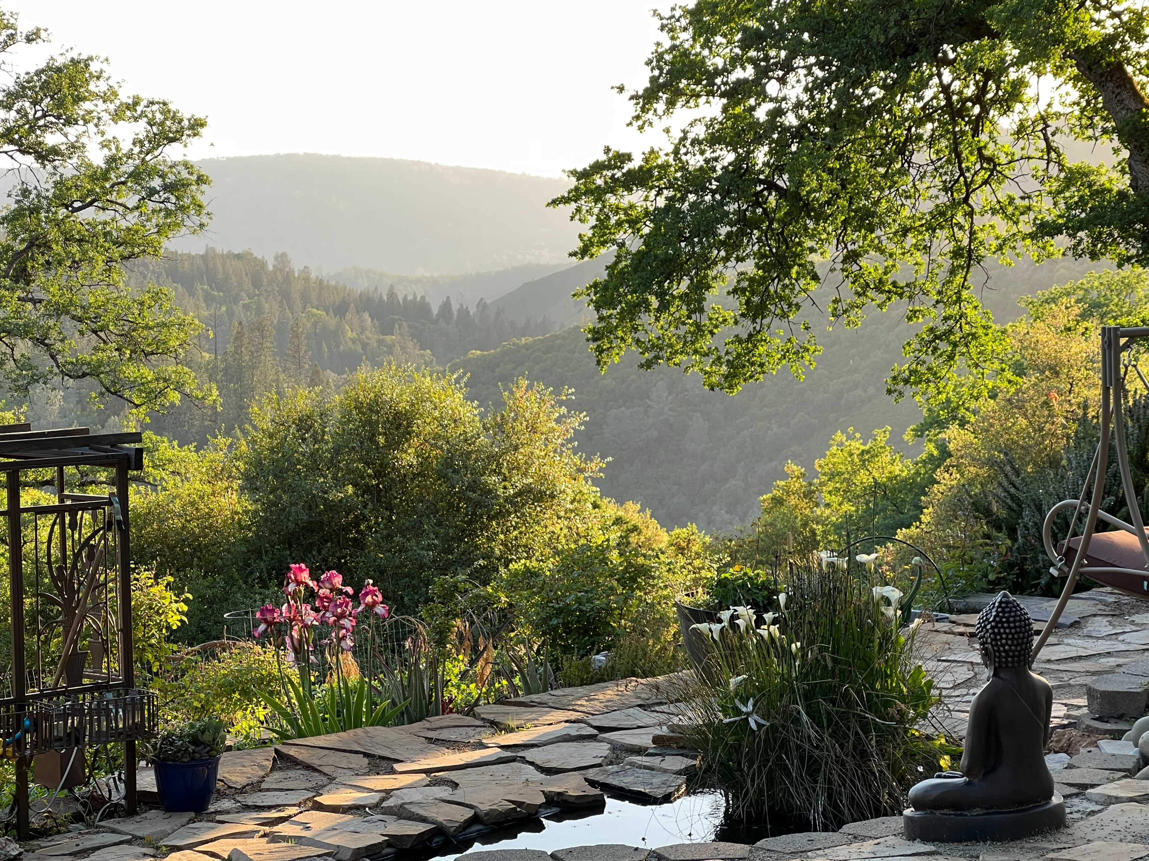 A stone pathway leads to a tranquil garden with a seated statue, overlooking lush green hills and flowers in bloom.