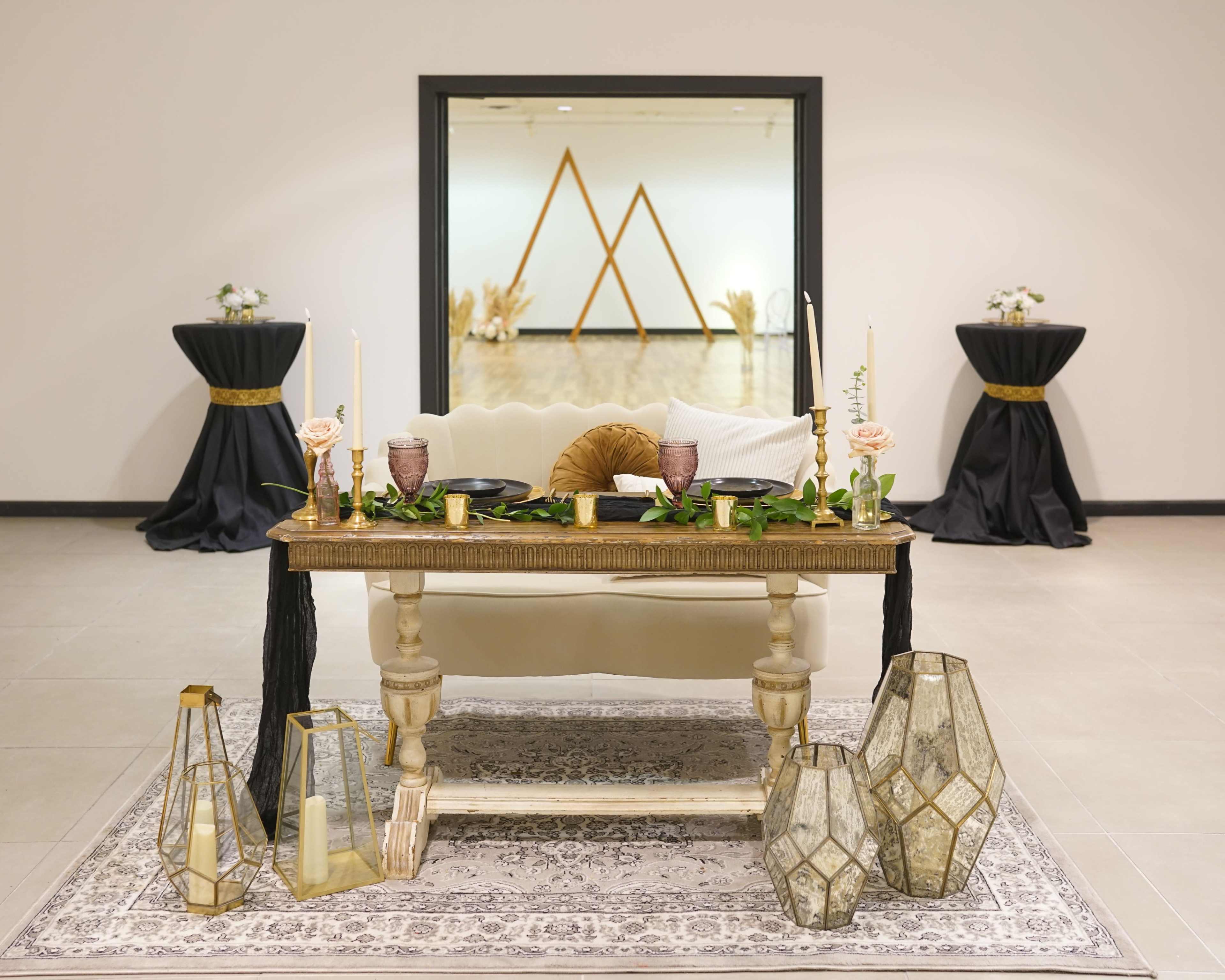A decorated table with candles and floral arrangements is set in front of a mirror reflecting two black-draped tables in a spacious room.