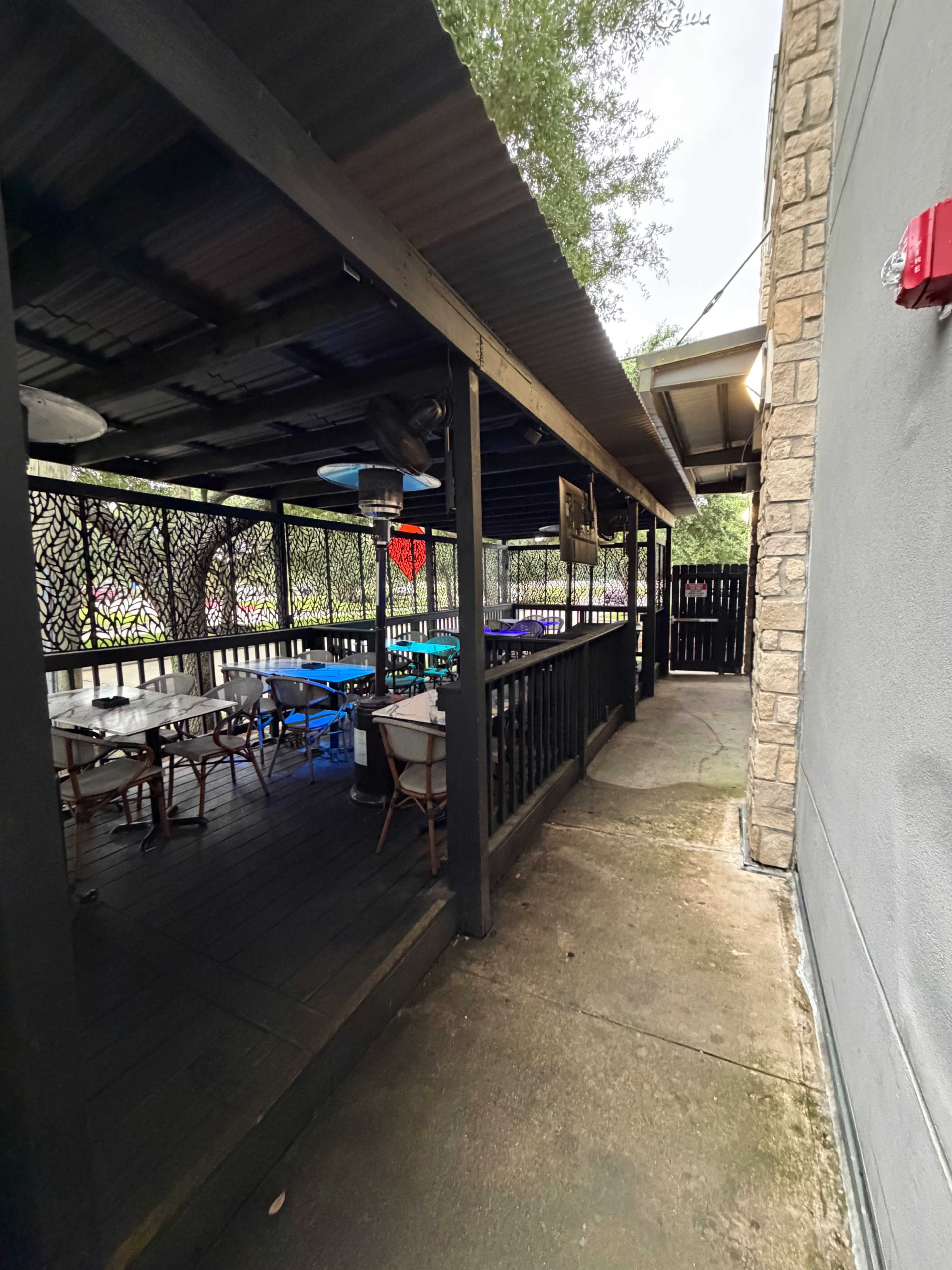 The image shows a covered outdoor seating area of a restaurant with wooden tables and chairs arranged along a walkway next to a stone wall.