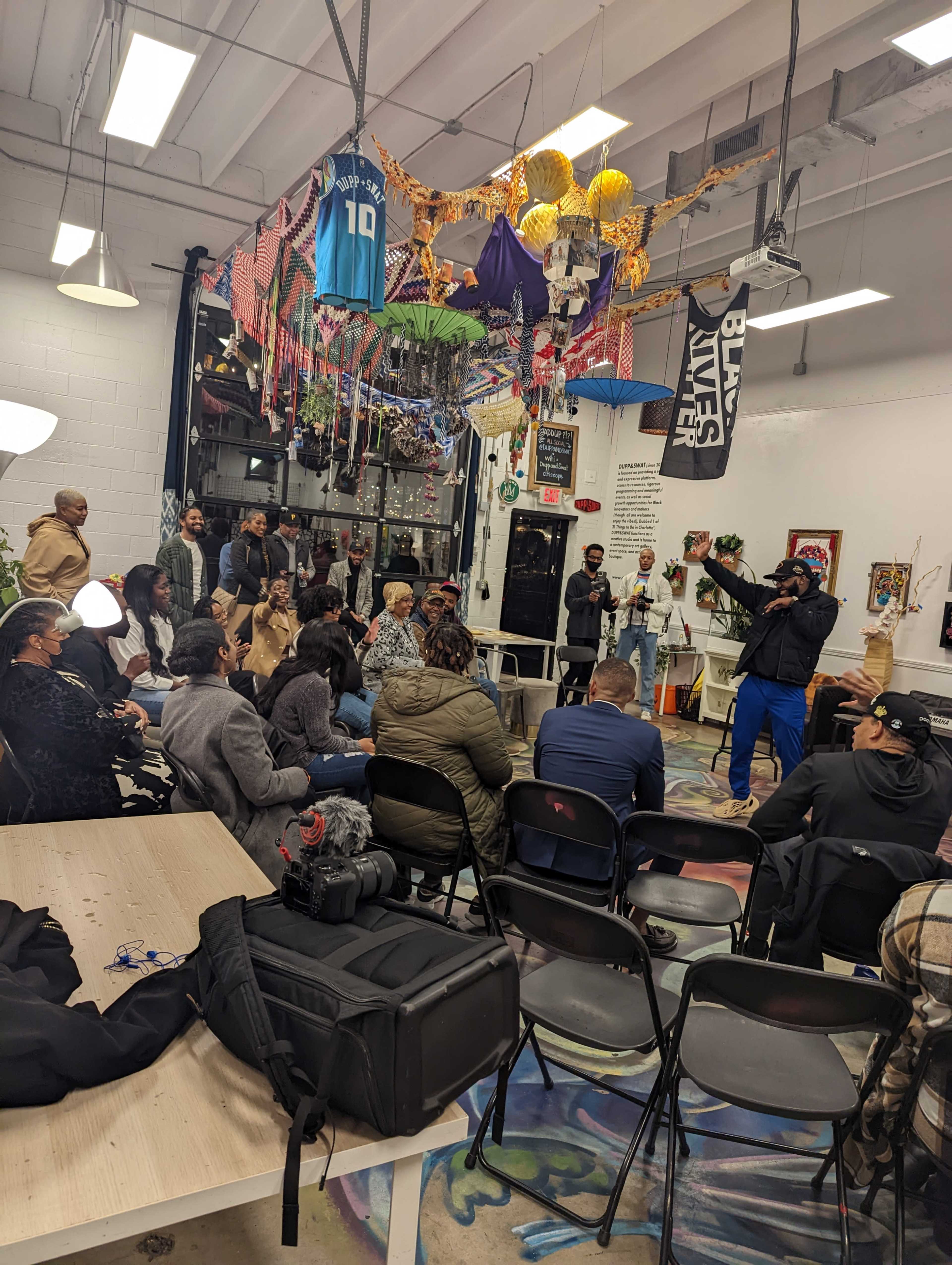 A diverse group of people is gathered in an art space, watching a speaker who is gesturing while standing in front of an audience seated at tables.