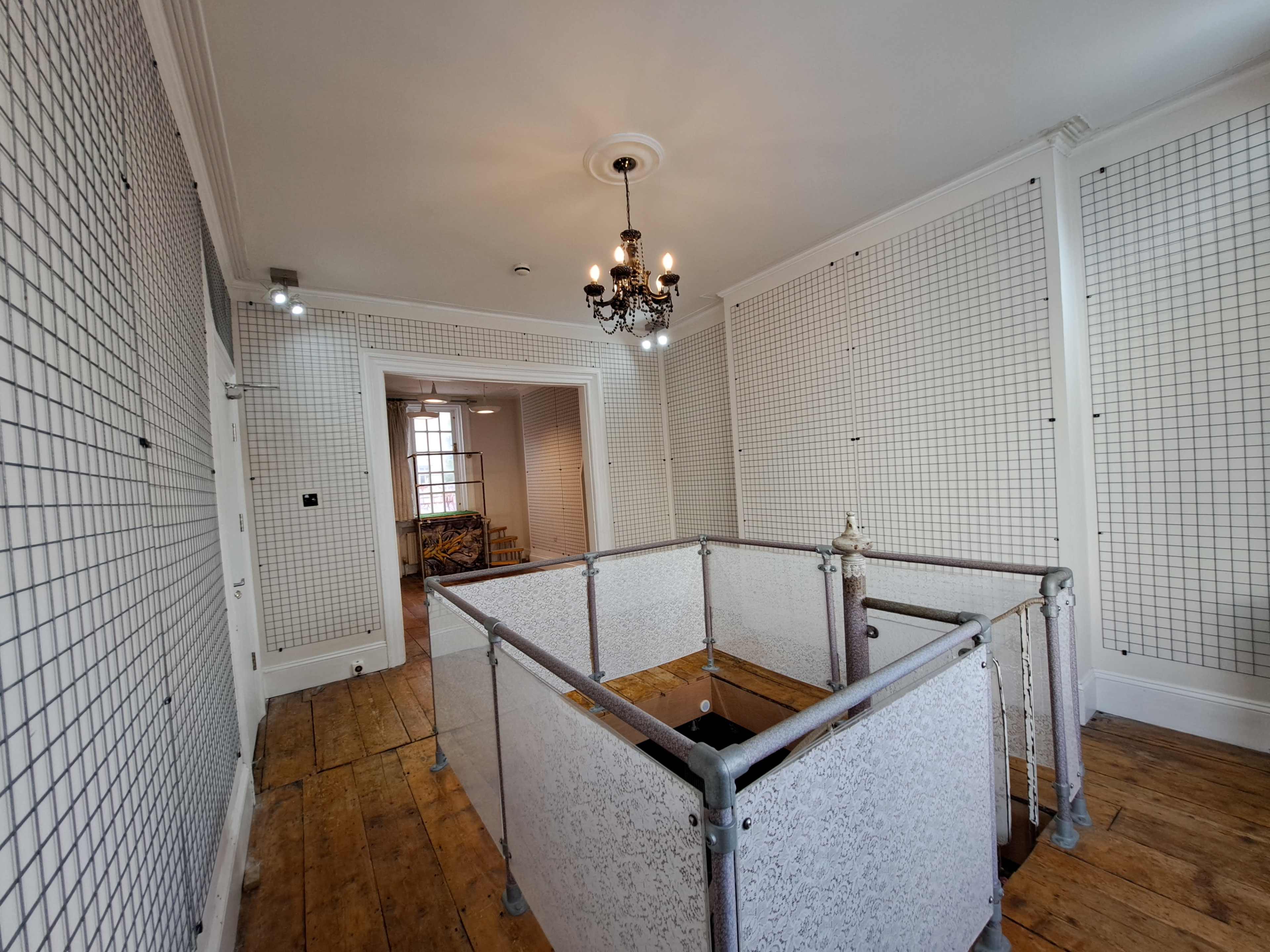 A spacious room with wooden flooring, grid-patterned wall panels, and a decorative chandelier, featuring a partial enclosure made of white mesh and metal.