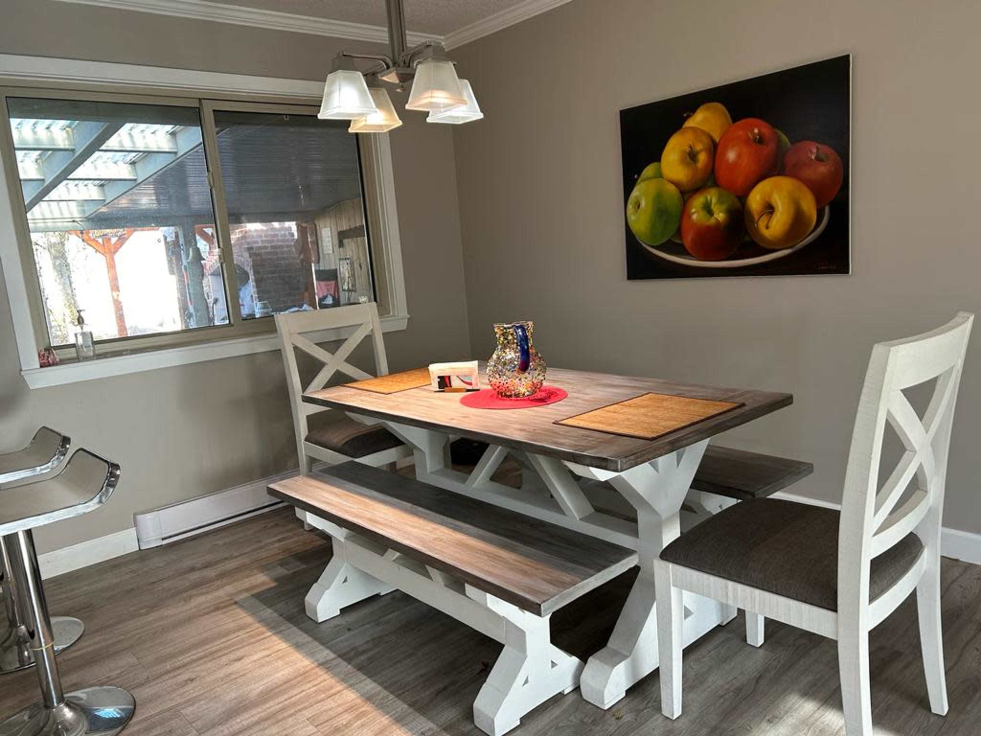 A dining area features a wooden table with benches and six chairs, illuminated by pendant lights, with a colorful fruit painting on the wall.