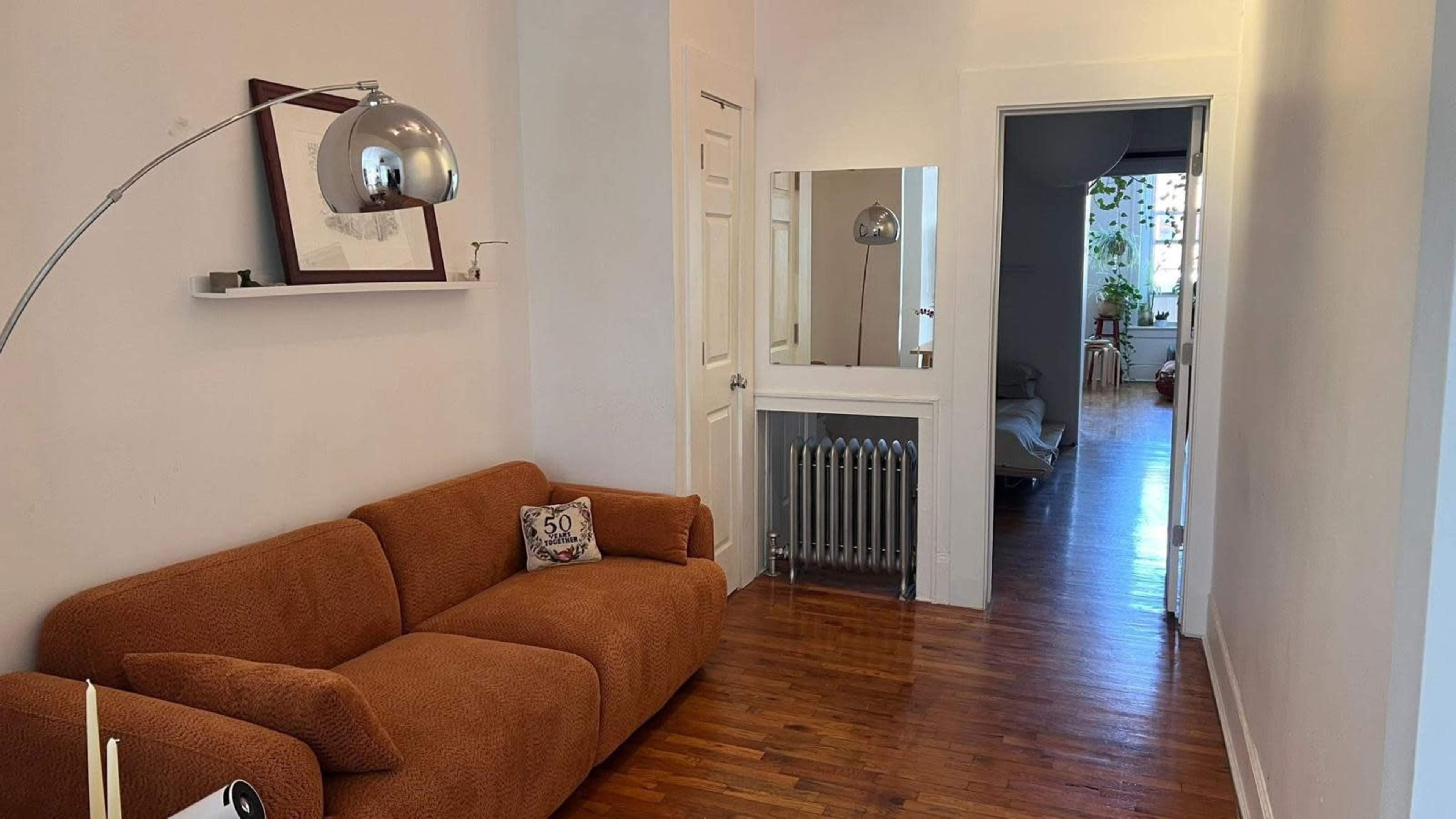 The image shows a living space with a brown couch, a curved floor lamp, and a doorway leading to another room, along with a mirror and a radiator in the hallway.