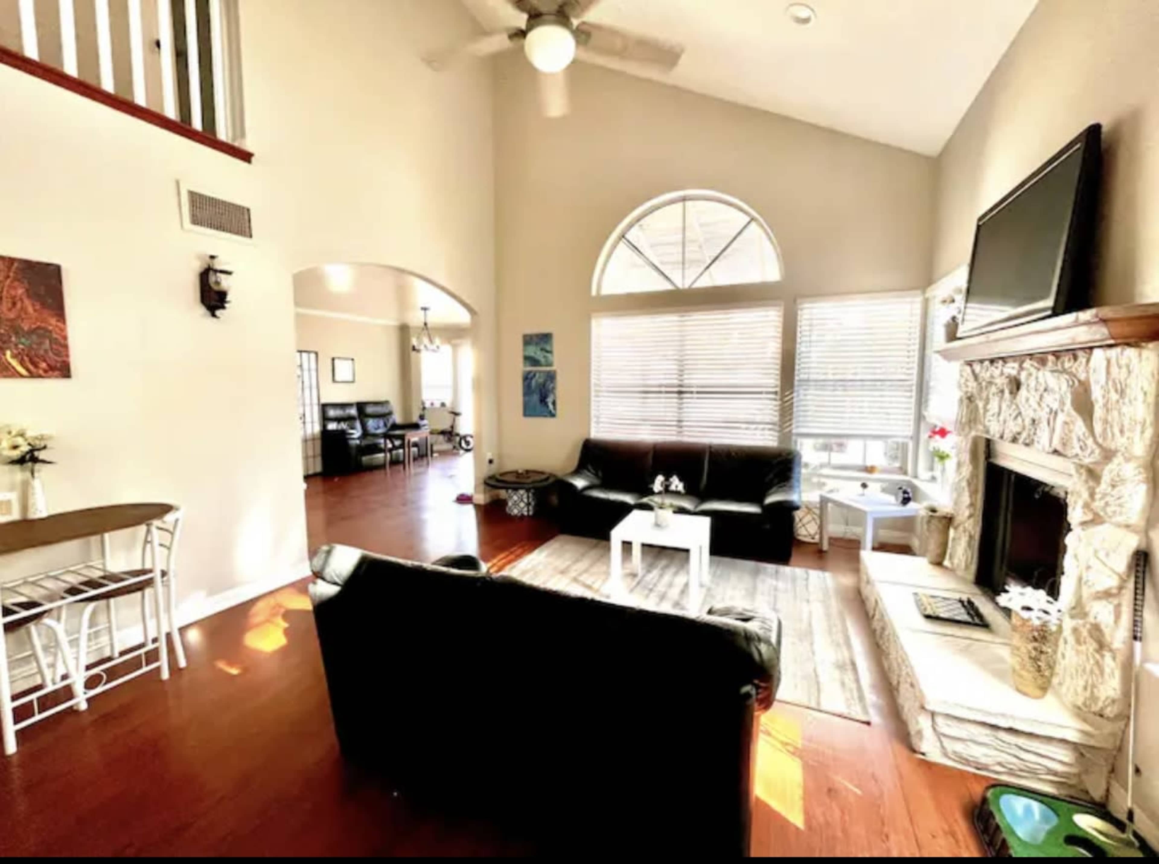 A spacious living room featuring a black leather sofa, a stone fireplace, and large windows that let in natural light.