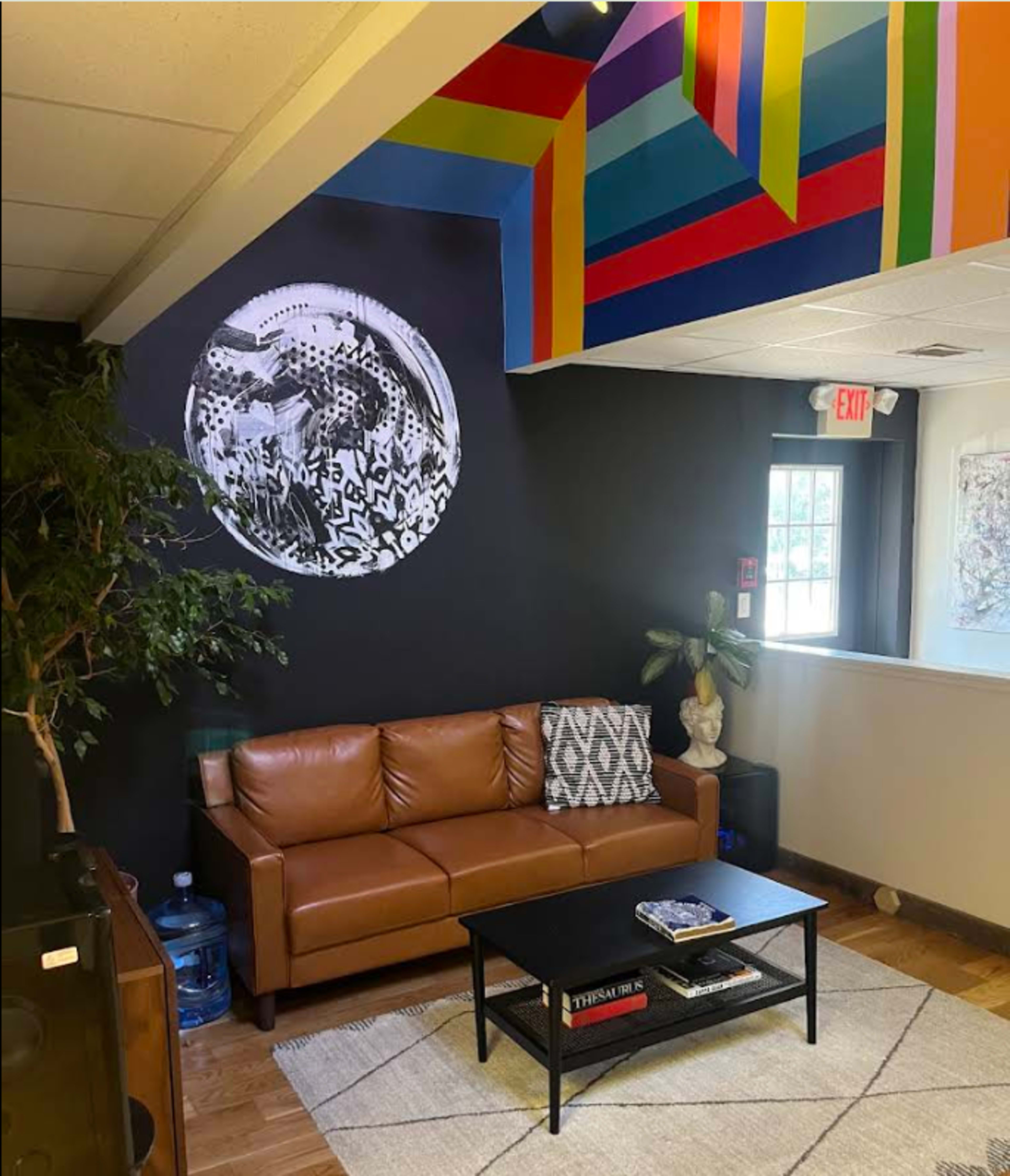 The image shows a modern lounge with a brown leather couch, a black accent wall featuring abstract art, and a colorful ceiling design.