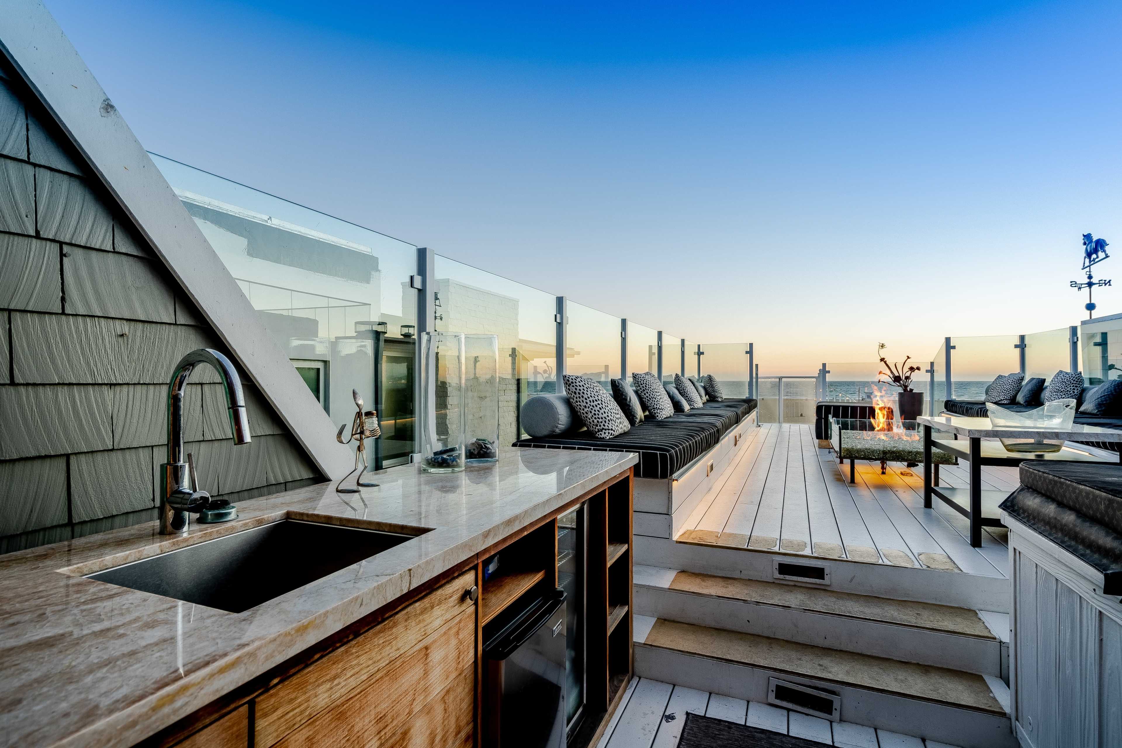 A rooftop terrace with seating areas, a small kitchen sink, and a fire pit against a backdrop of a sunset sky.