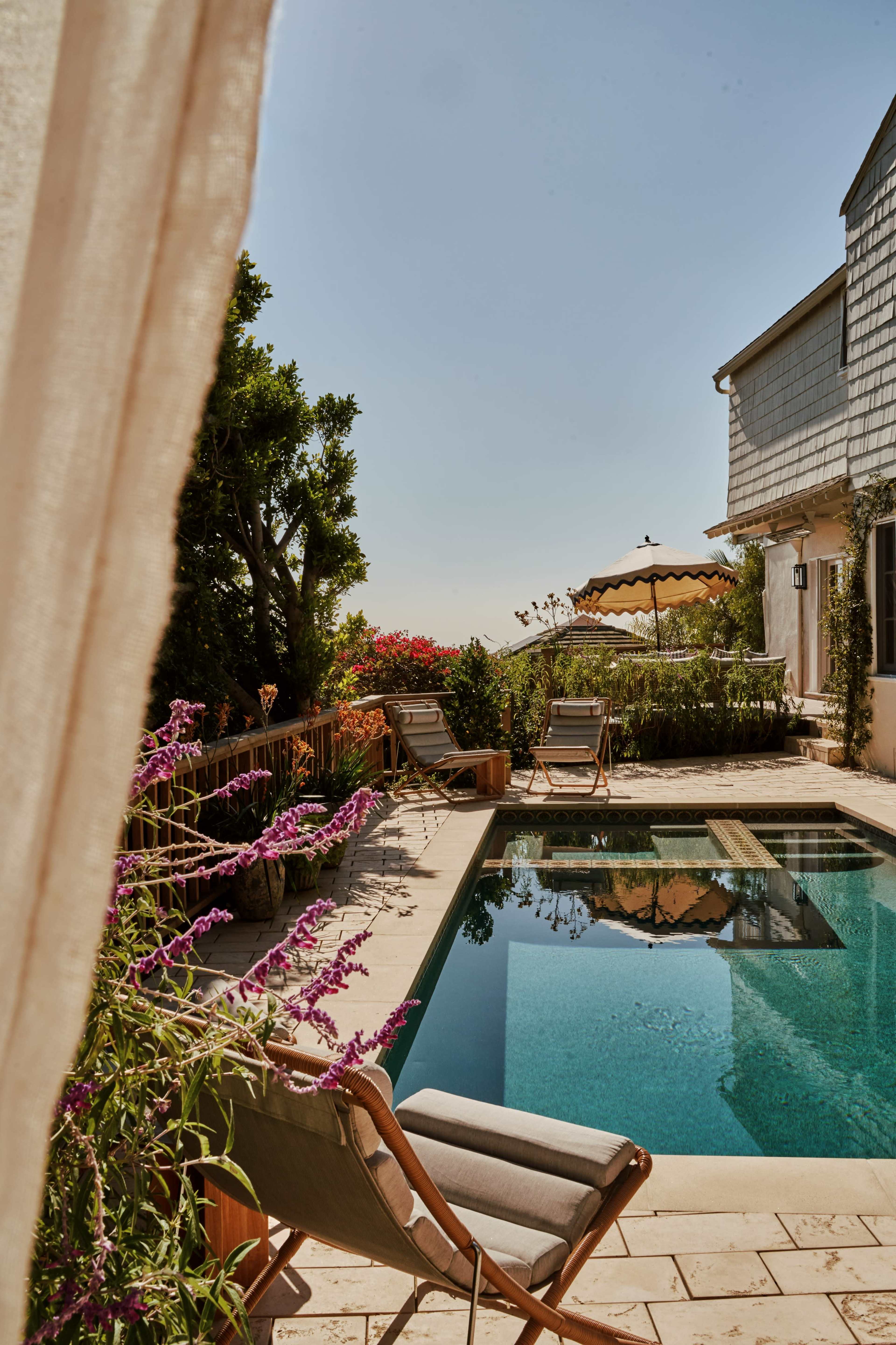 The image depicts a serene outdoor setting featuring a swimming pool surrounded by lounge chairs, lush greenery, and a clear blue sky.