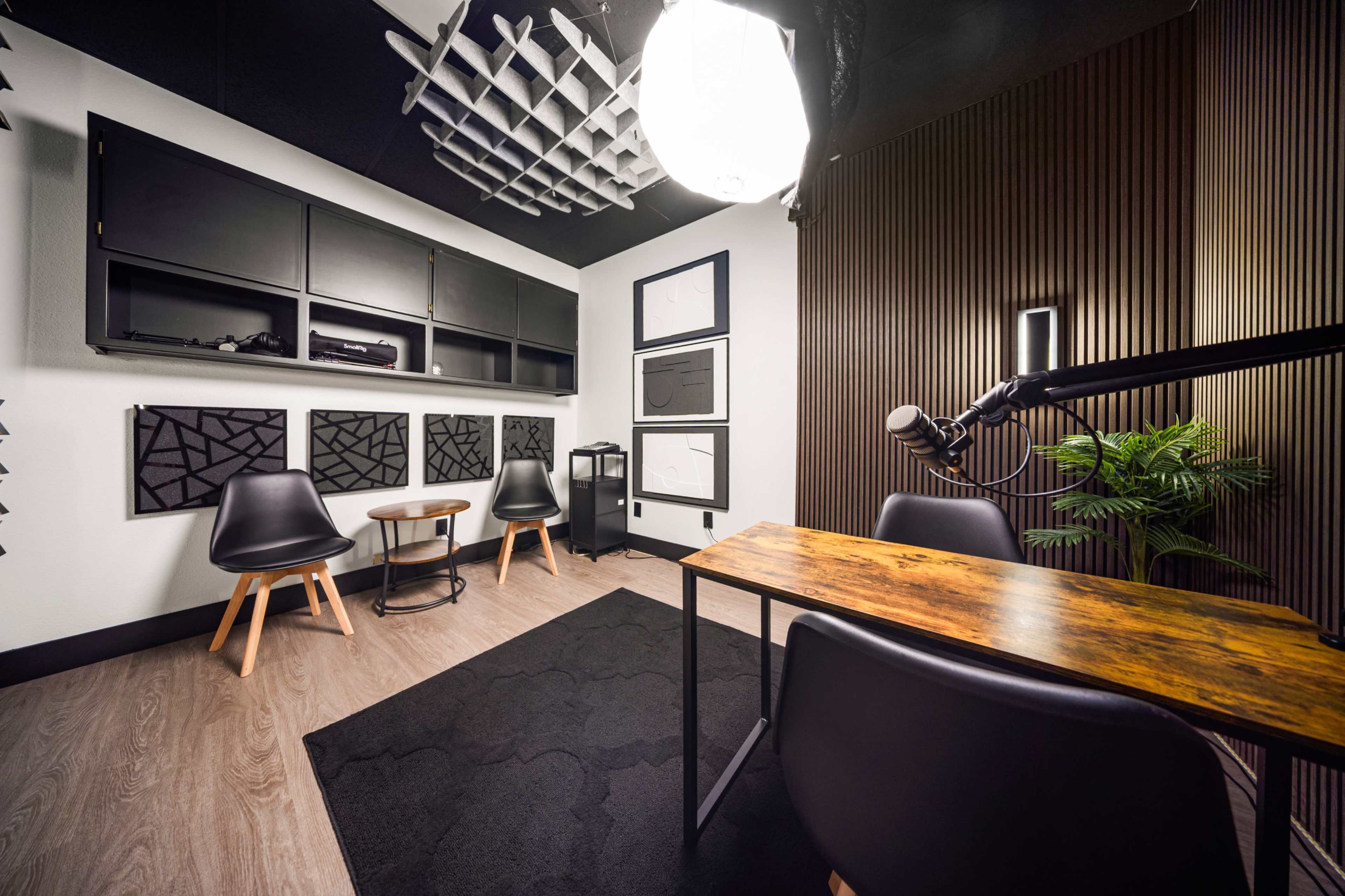 The image shows a stylish, modern recording studio with a small round table, two chairs, a desk, and soundproofing panels on the walls.
