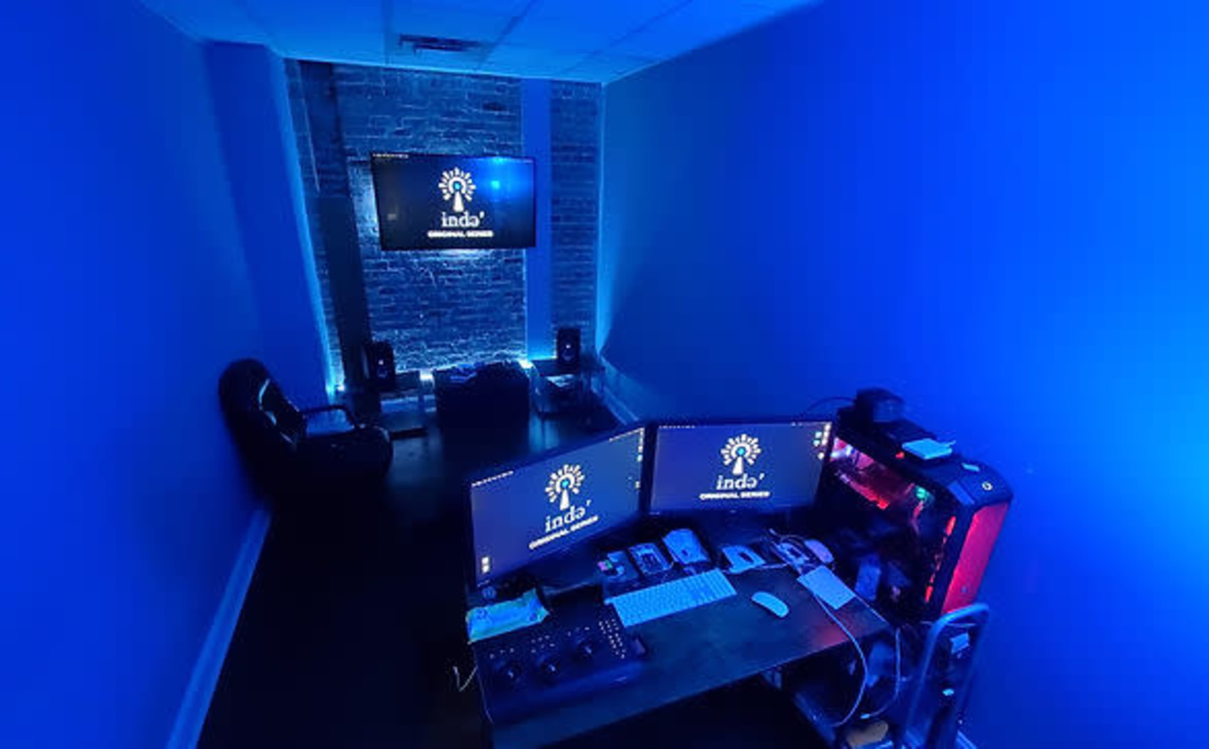 A dimly lit office space with blue lighting, featuring two computer monitors, a desk with various devices, and a television on the wall.