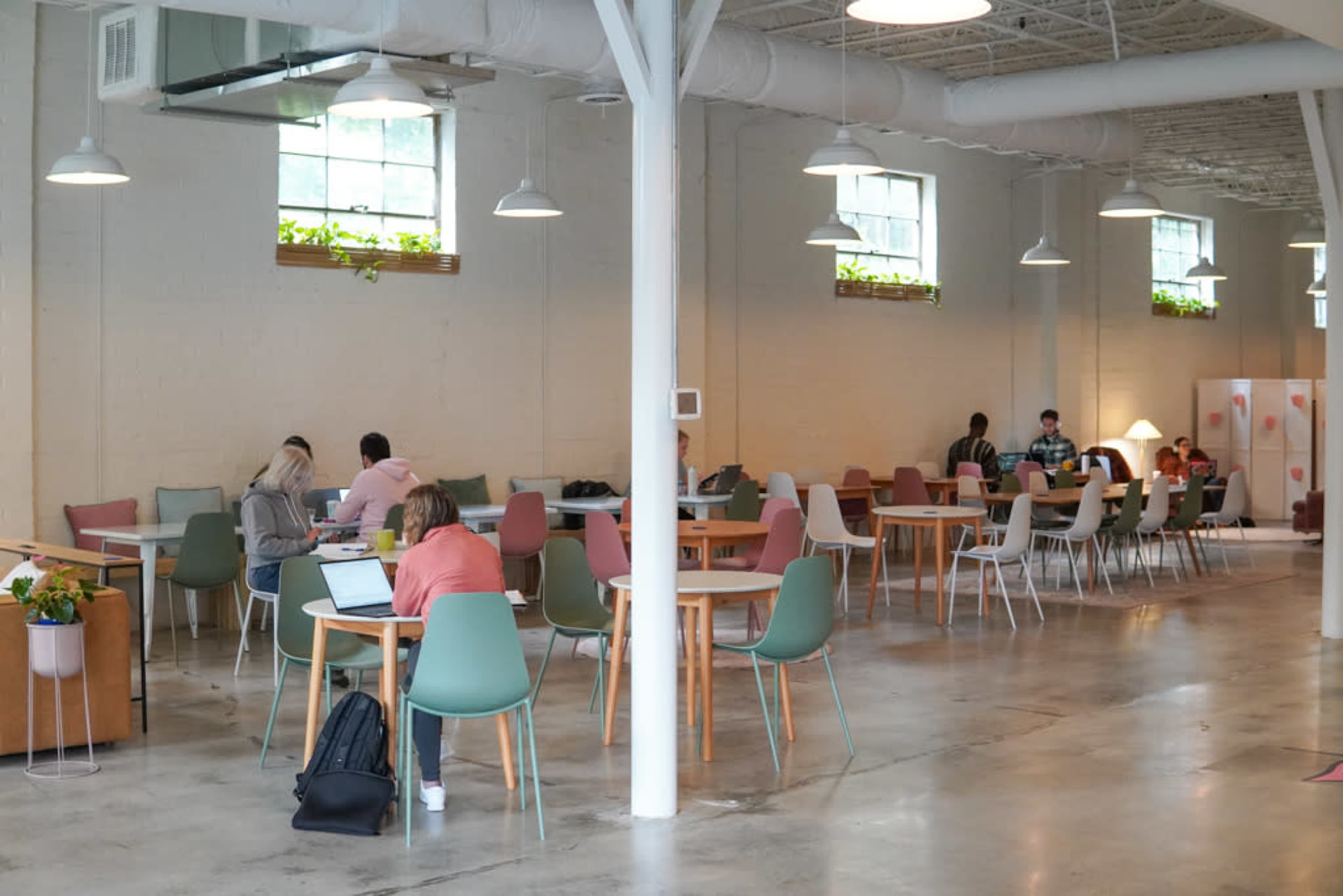 A spacious co-working area with various tables and chairs, where people are engaged in different activities such as working on laptops and conversing.