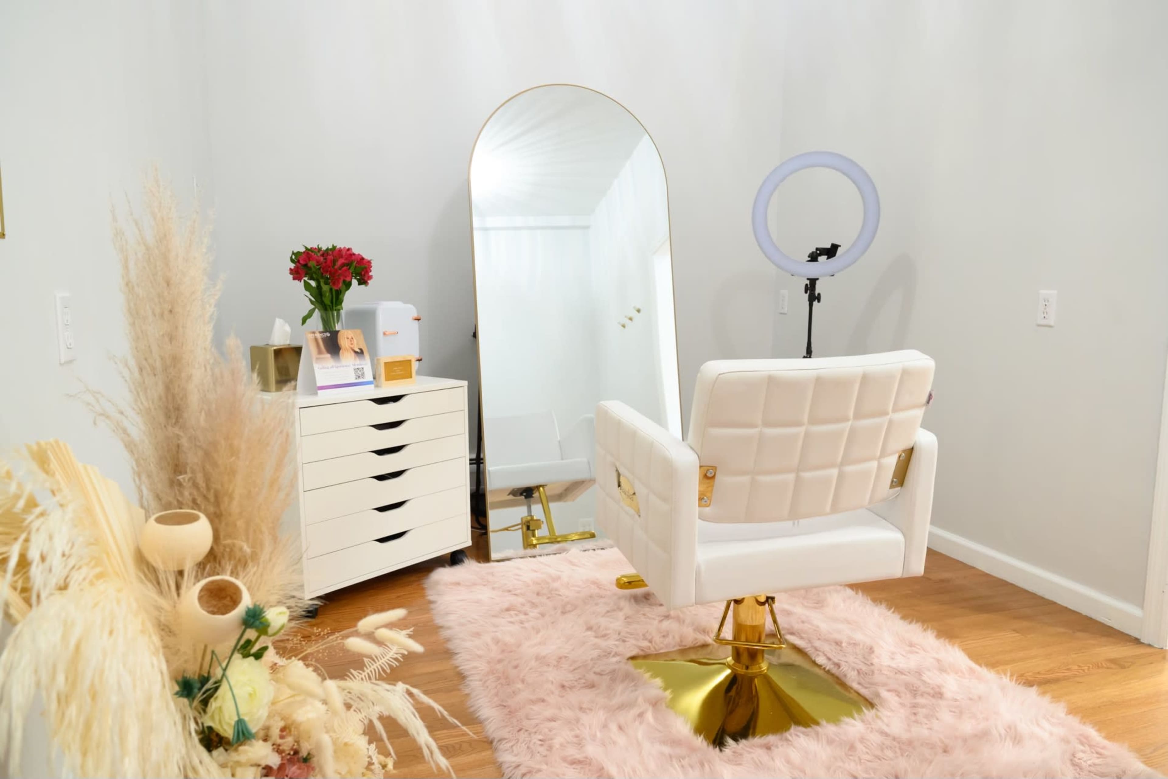 The image shows a modern beauty salon setup featuring a white styling chair, a large mirror, a ring light, and a storage unit with drawers, all arranged on a soft pink rug.