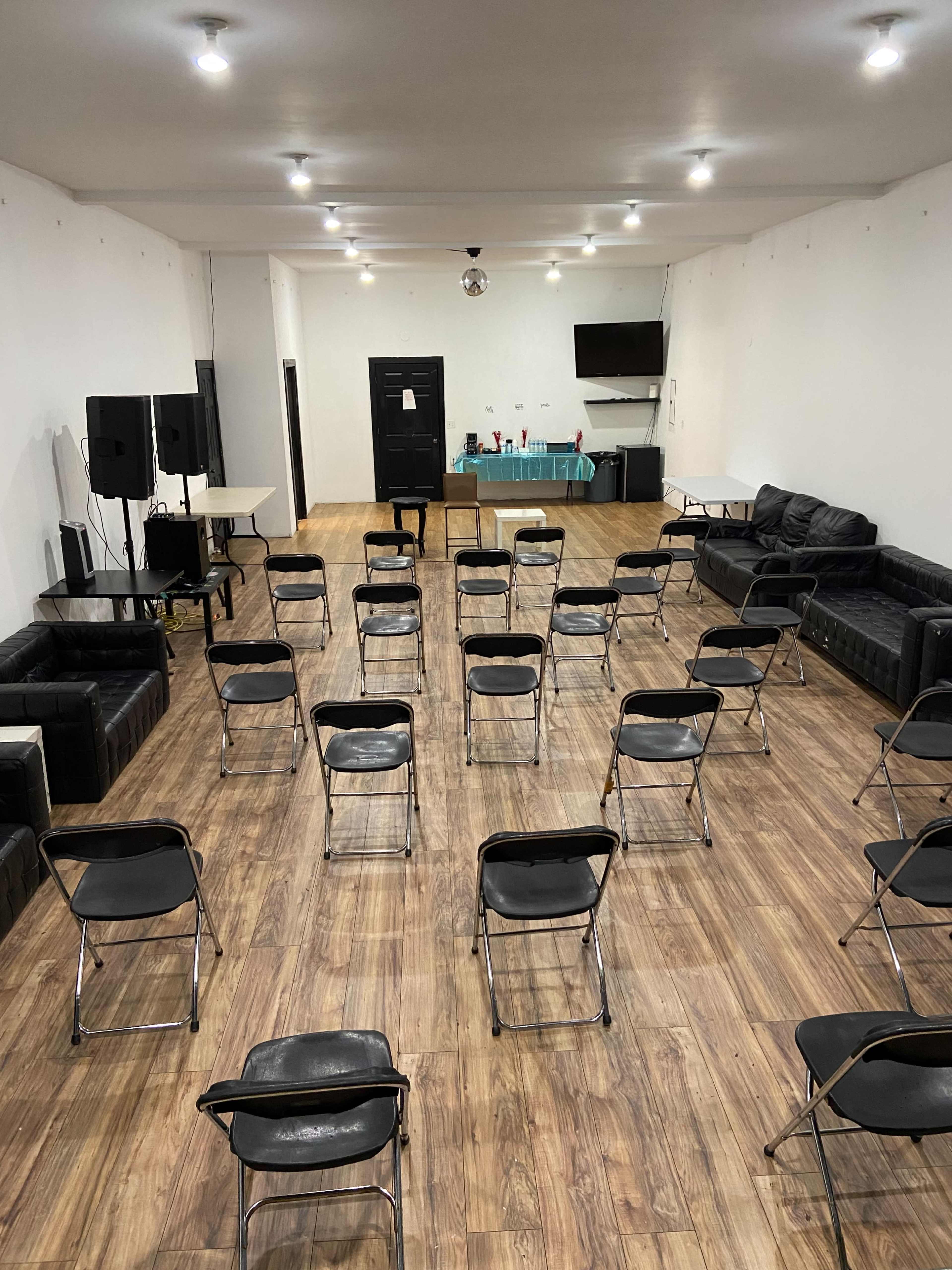 A spacious indoor room with a wooden floor arranged with metal chairs set in rows facing a black door and a table in the background.