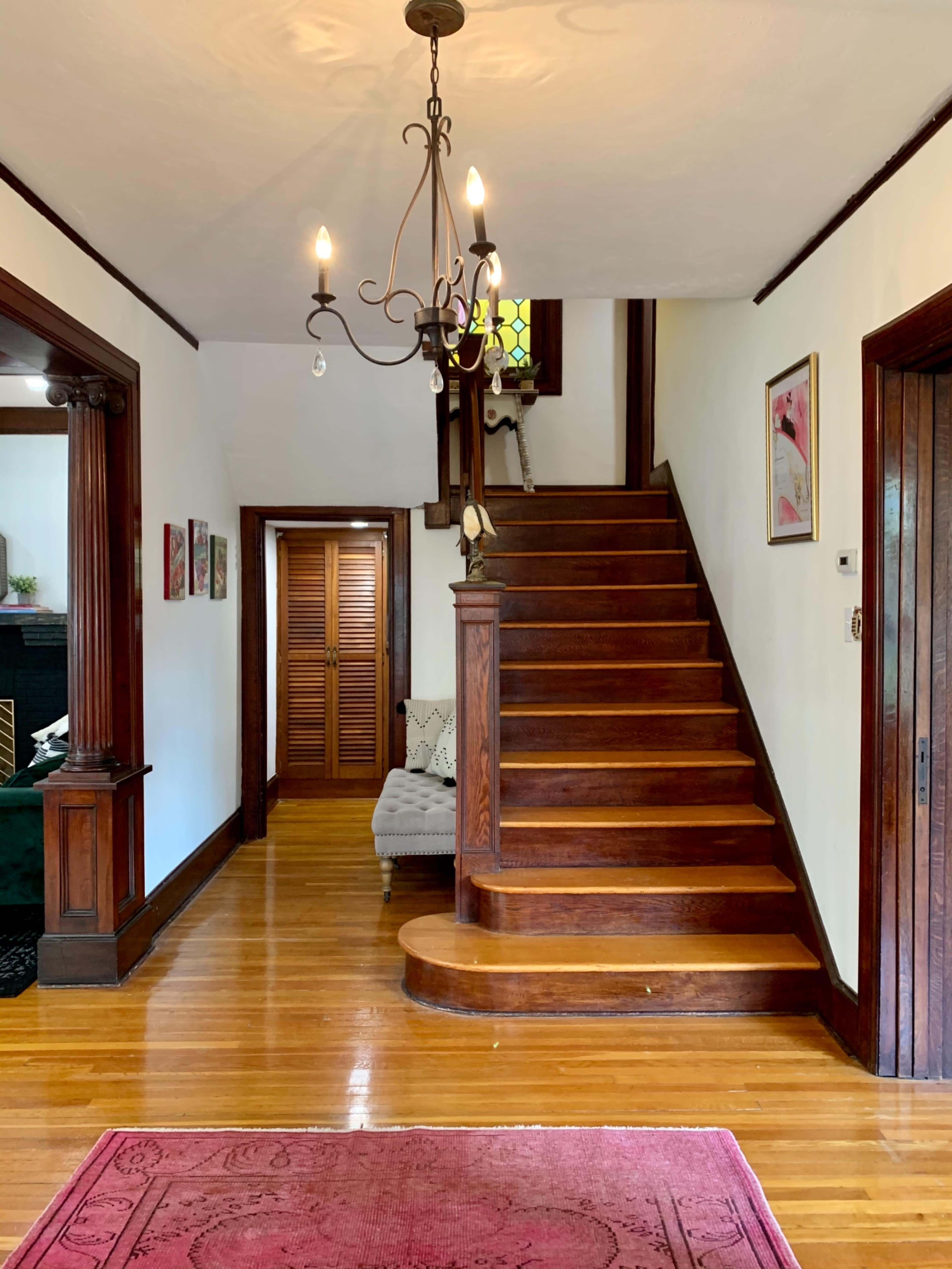 The image shows a hallway with a staircase leading to an upper floor, featuring wooden steps and a chandelier, along with a cozy seating area and doorways to adjacent rooms.