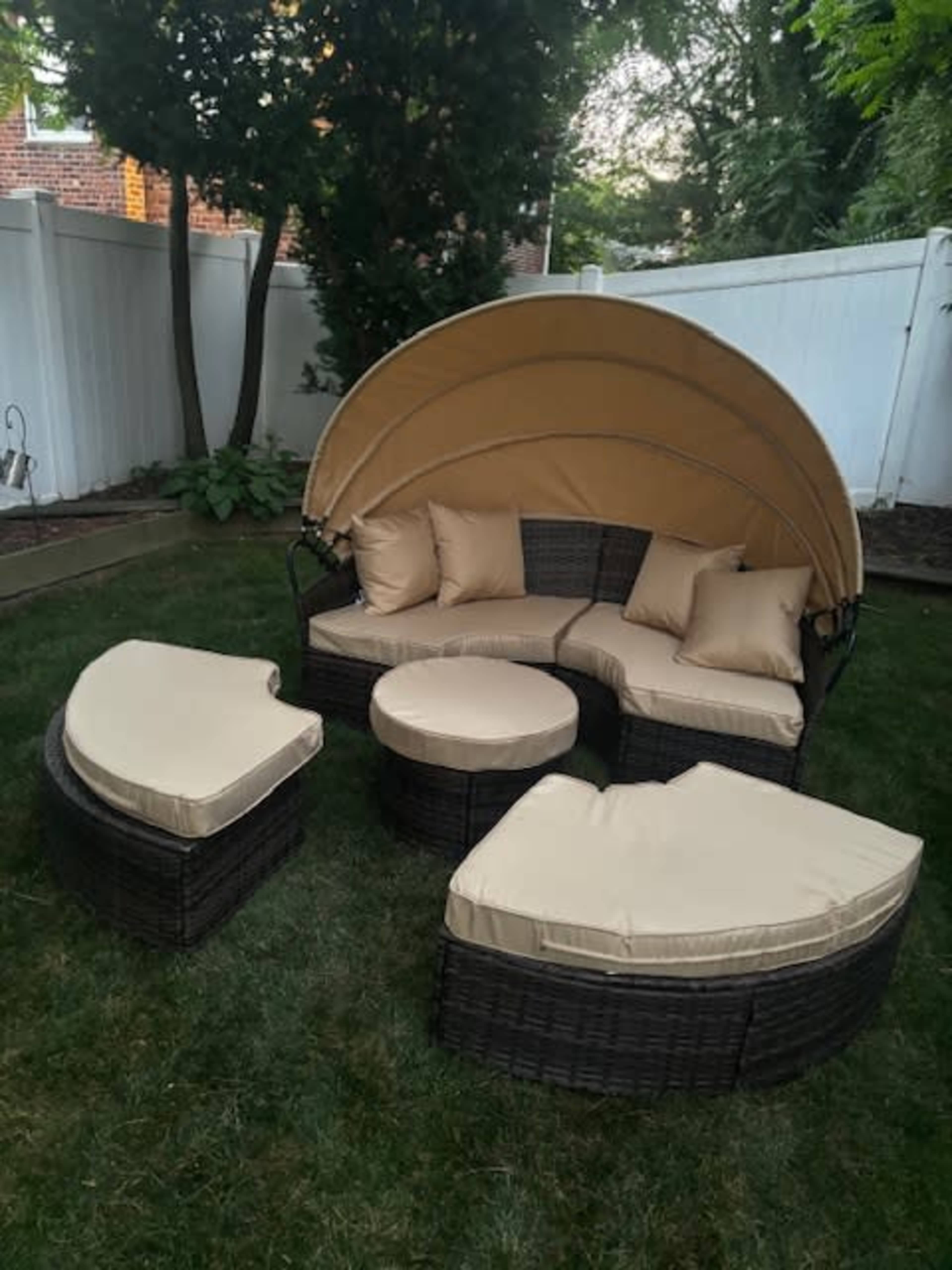 A curved outdoor seating arrangement with a canopy and cushions is set on a grassy area surrounded by a white fence.