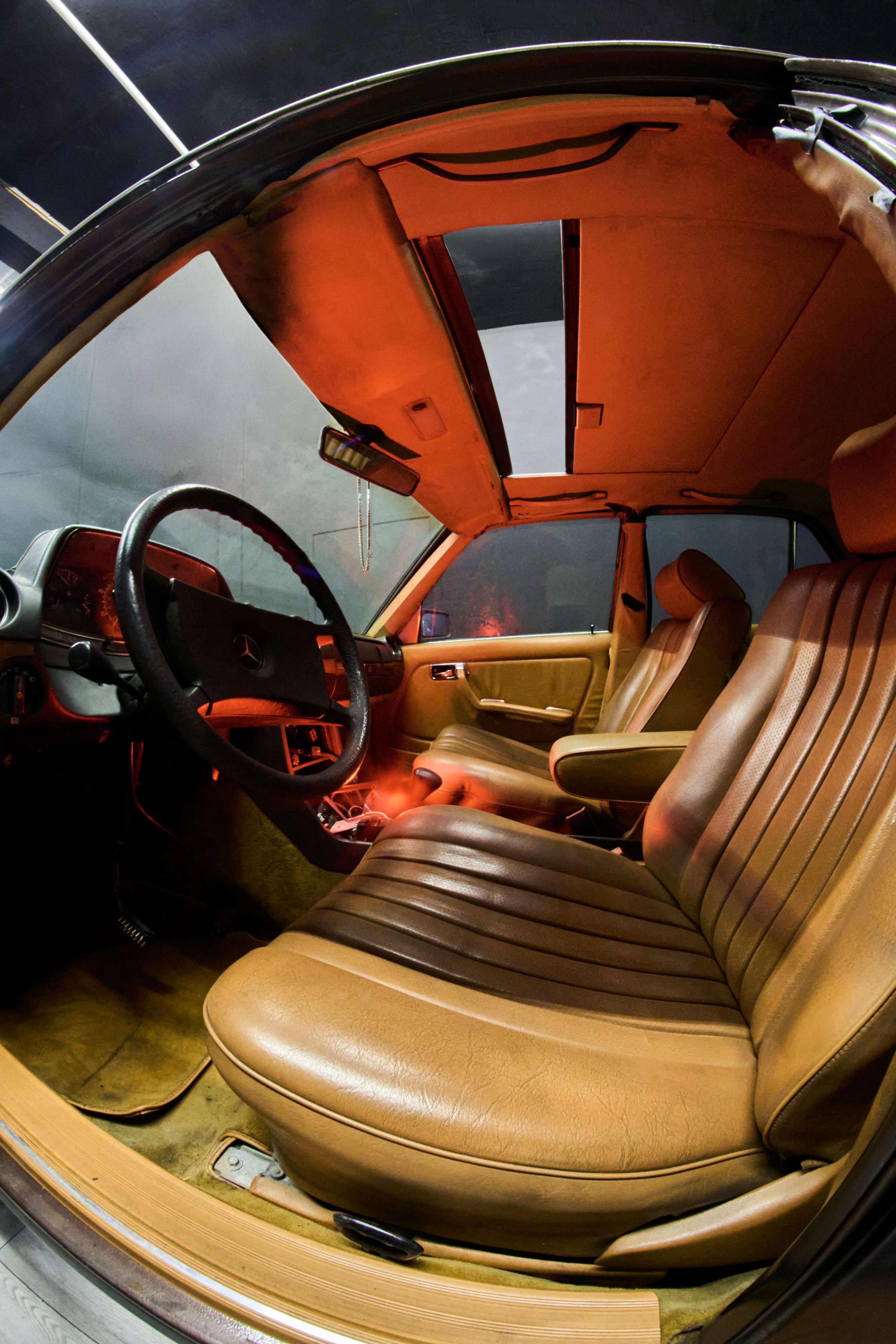 The interior of a classic Mercedes-Benz car features brown leather seating and a dashboard with various controls, illuminated by a soft red light.