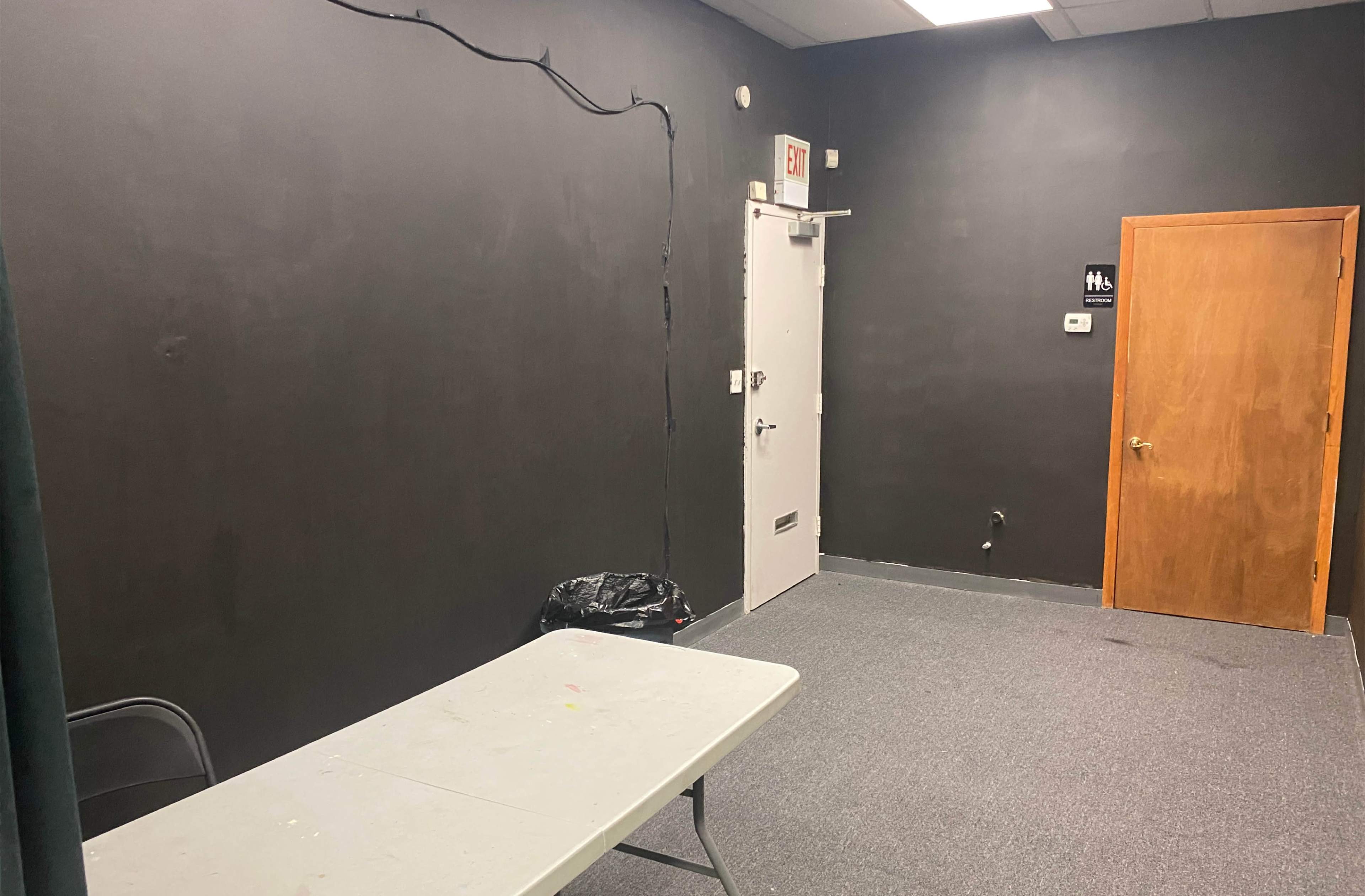 The image shows a small room with a dark gray wall, a wooden door, a table, a chair, and a wastebasket in the corner.